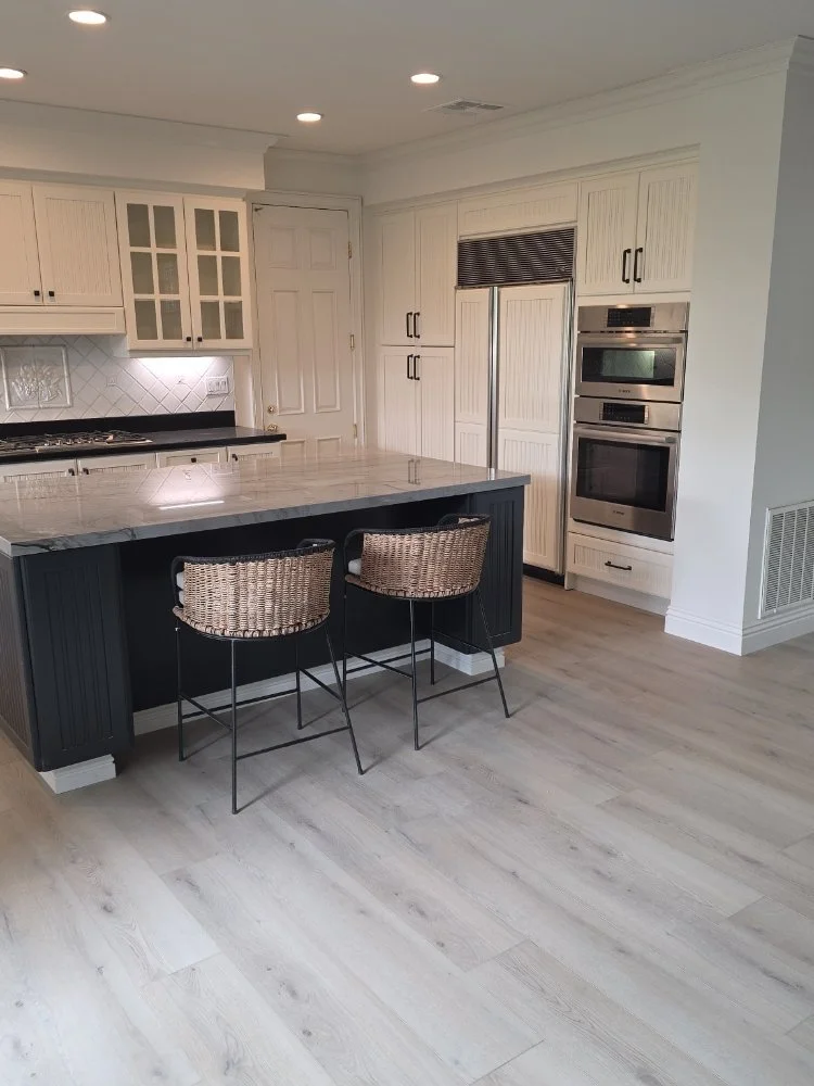 Modern kitchen with white cabinets, stainless steel double oven, marble-topped island with black base, two woven bar stools, and light wood flooring.