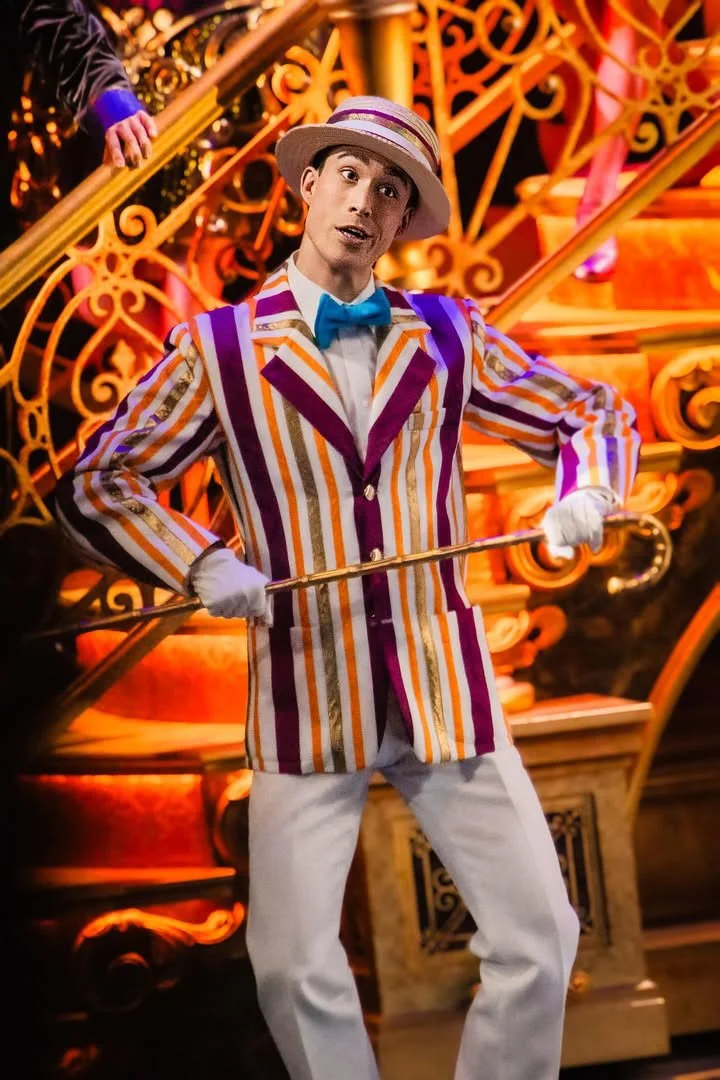 A man dressed in colorful, striped jacket with purple, orange, and white, wearing a white hat with purple and orange stripes, blue bow tie, white gloves, and white pants, holding a cane, standing in front of an ornate, gold and orange staircase with intricate detailing.