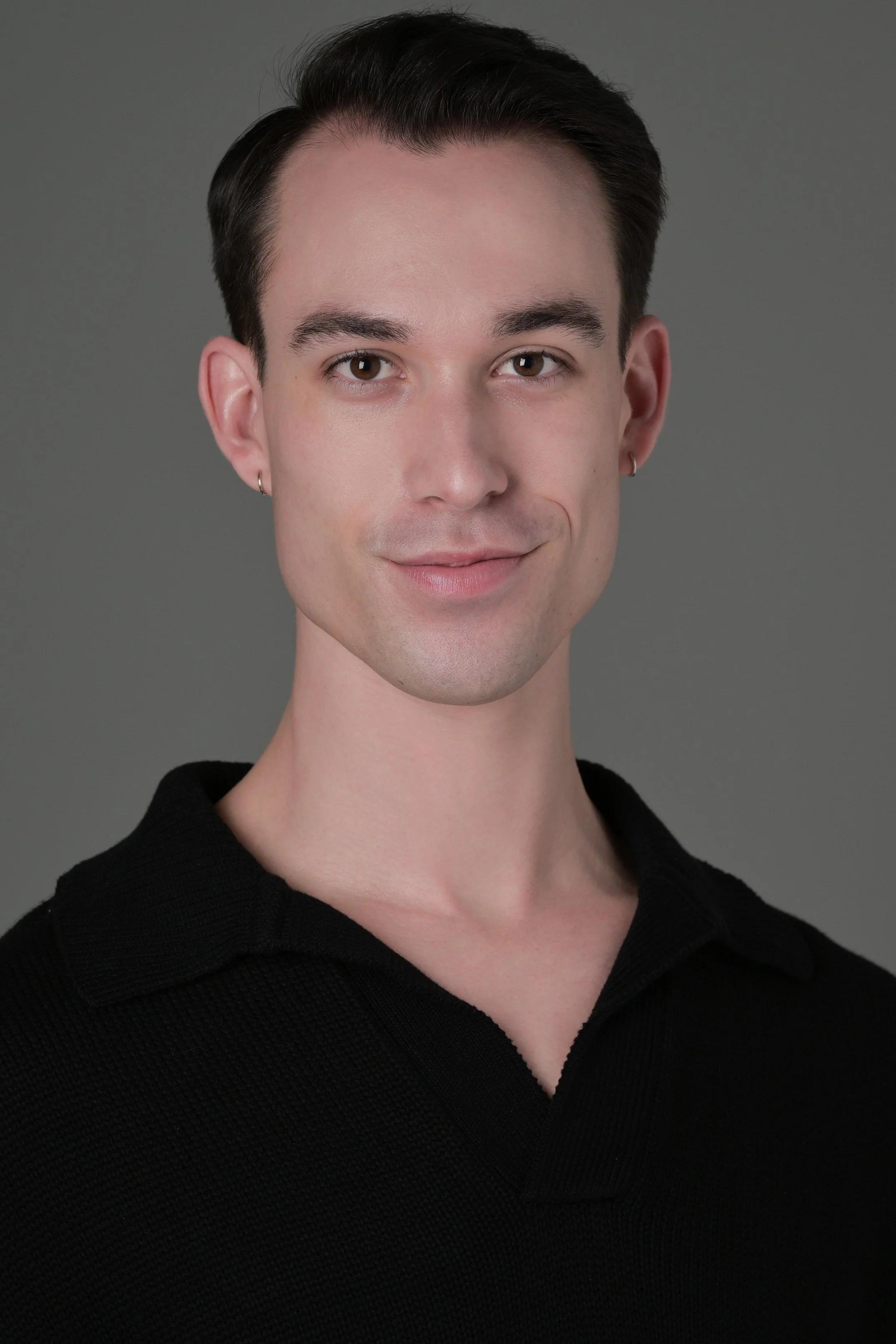 Close-up of a young man with short dark hair, light skin, earrings, wearing a black shirt, smiling slightly, against a gray background.