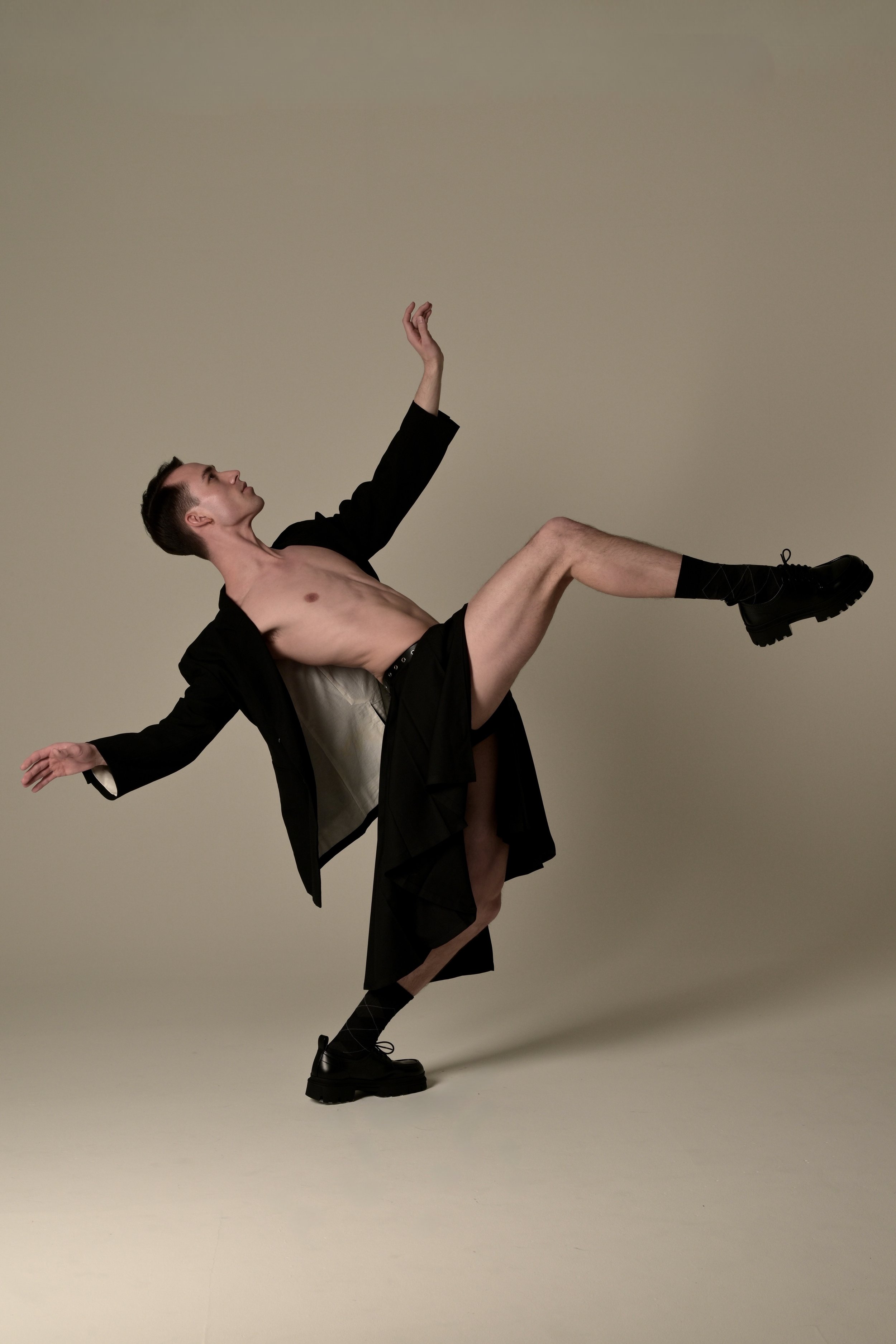 A shirtless man wearing black shorts, a black jacket, and black boots is balancing on one foot with the other leg lifted, in a dance pose against a plain background.