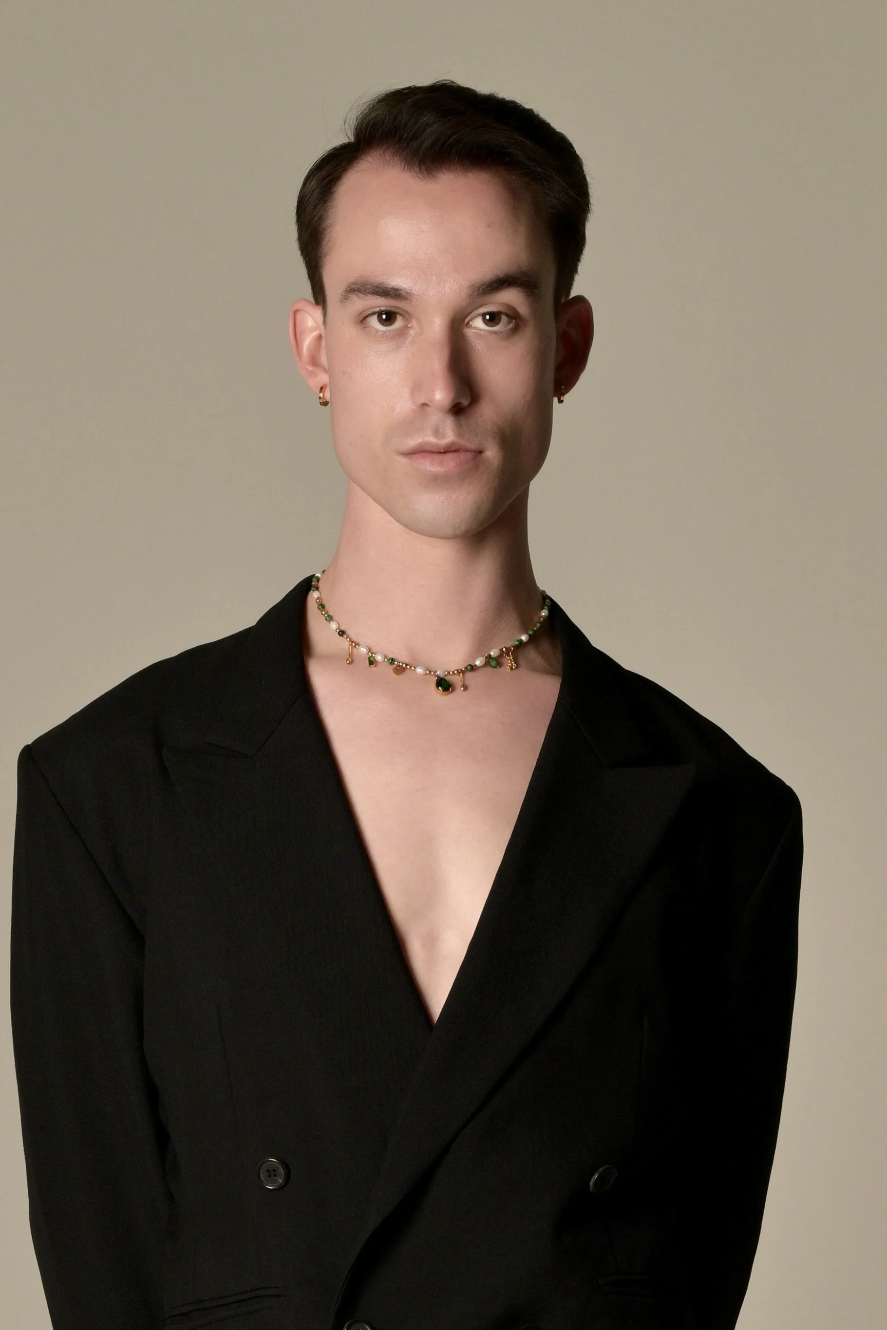 A person with short, dark hair styled upward, wearing gold hoop earrings, a multicolored beaded necklace, and a black blazer with button details, standing against a plain gray background.