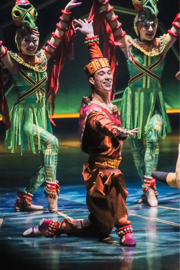 Performer in traditional African costume, kneeling with arms extended, surrounded by performers in colorful tribal attire and masks, stage with dramatic lighting.