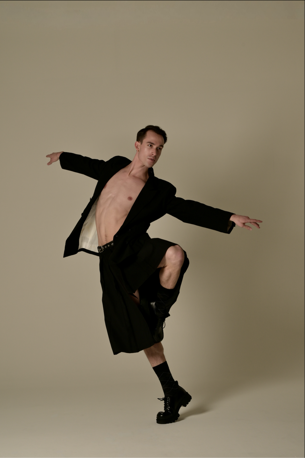 A person with a bare chest, wearing a black blazer, black shorts, black shoes, and socks, is balancing on one leg with arms extended, against a plain beige background.