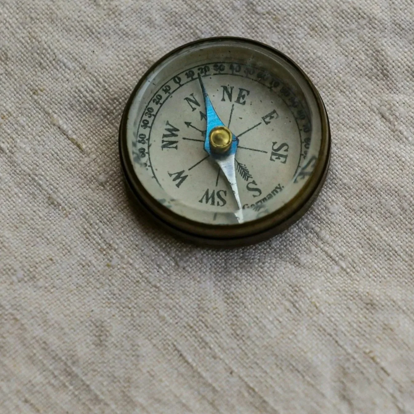 A vintage compass with a metallic casing and a white face, displaying directional markings and a blue arrow pointing north, resting on a textured beige fabric surface.