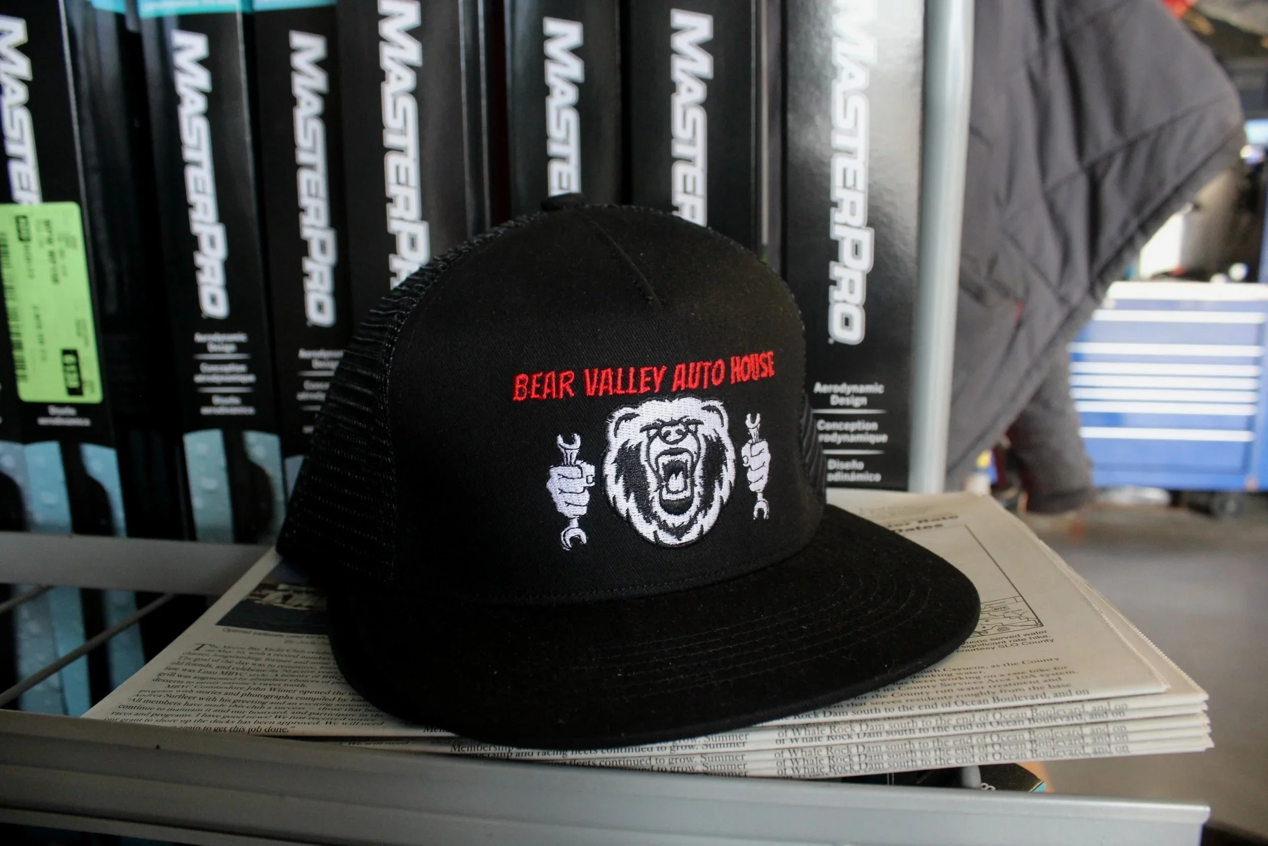 A black baseball cap with a bear logo and 'Bear Valley Auto House' embroidered in red, placed on top of a pile of papers on a white shelf in a store. In the background, black boxes with white labels and a person in a gray jacket are visible.