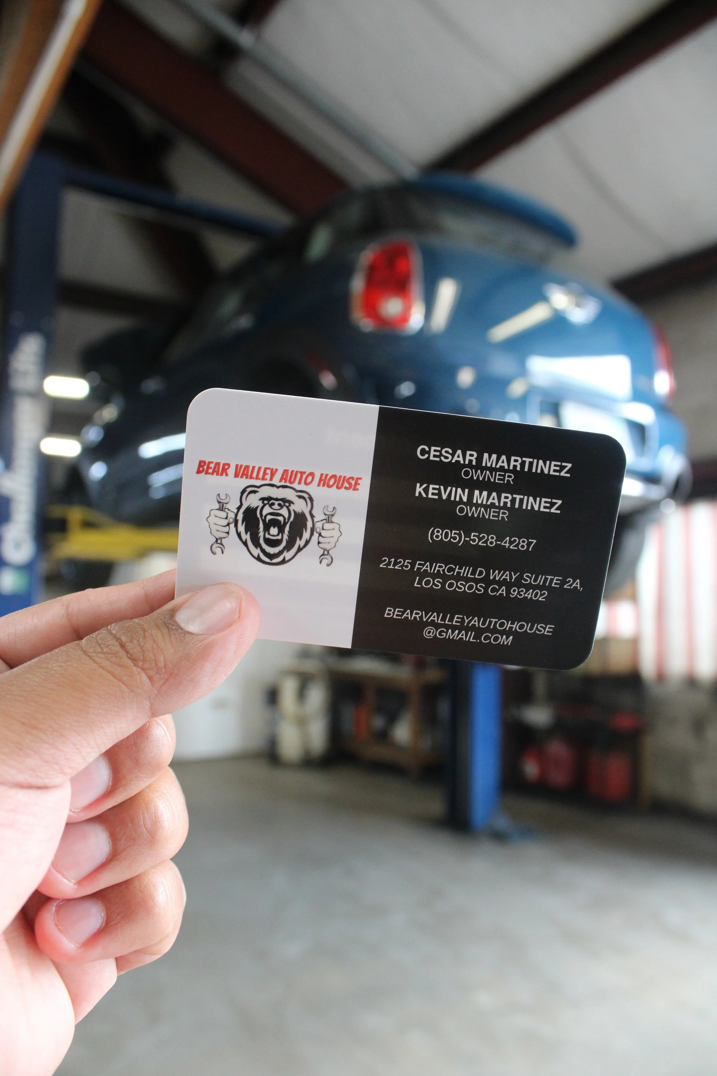 A person holding a business card for Bear Valley Auto House, with a blurred blue car on a lift in the background inside a garage or auto repair shop.