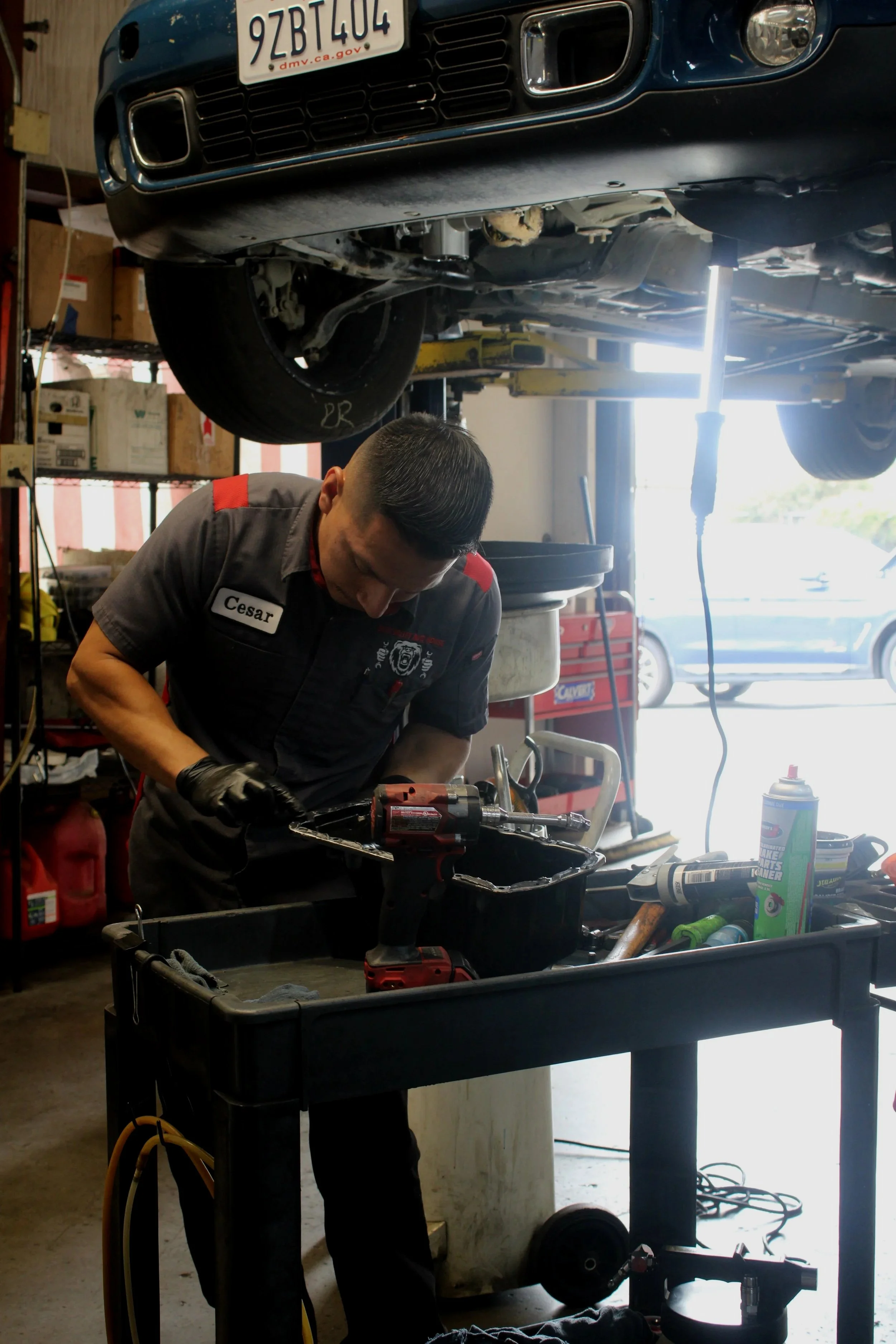 A mechanic working underneath a car lifted on a hydraulic lift in an auto repair shop. The car's front is visible with a license plate, and the mechanic is wearing a black work shirt with a name tag that reads 'Cesar.' Tools and automotive parts are scattered around.