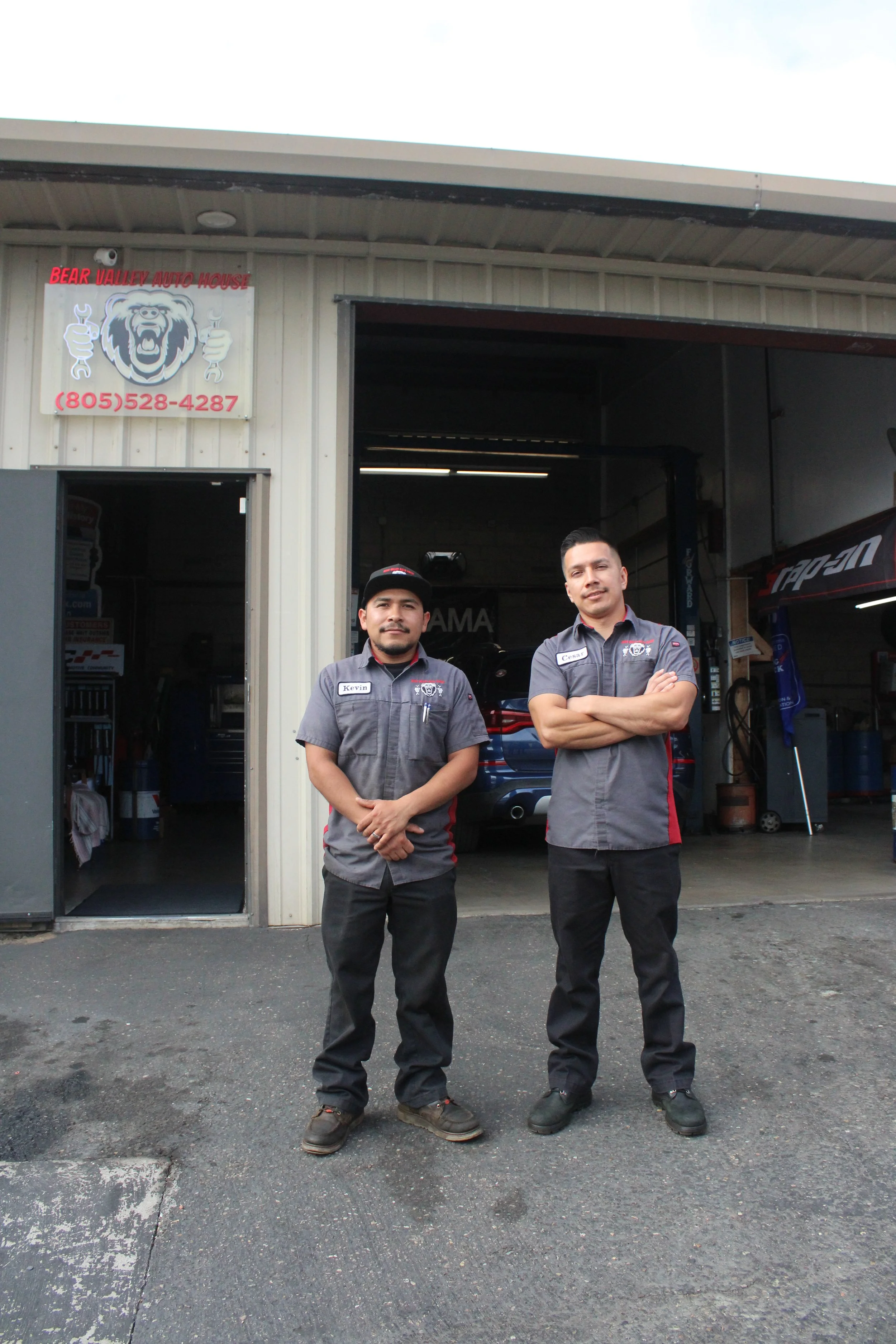 Two men wearing work uniforms standing outside automotive repair shop with open garage door. Sign on building reads 'Bear Valley Auto House' and includes a phone number.