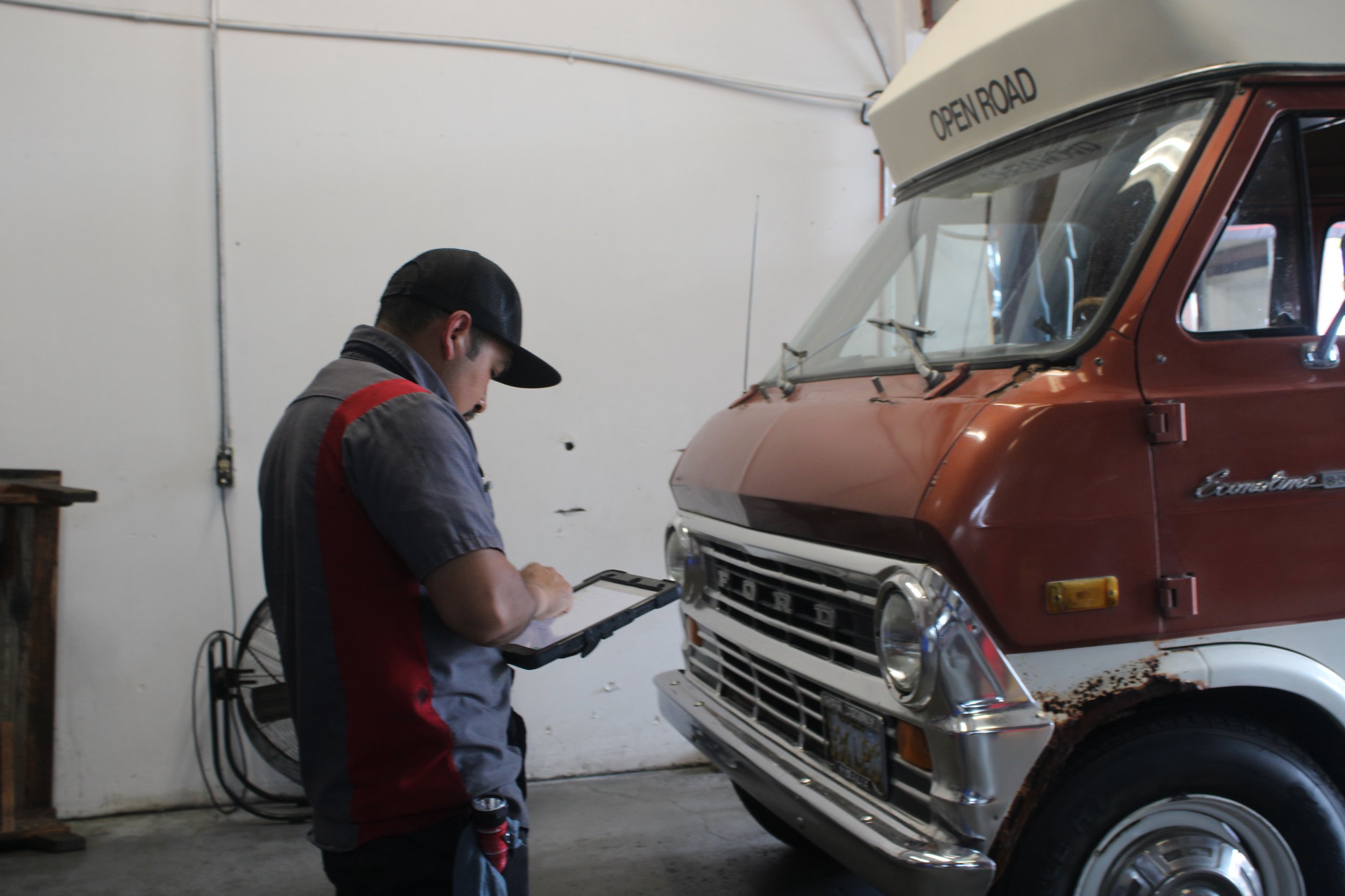 A mechanic working with a clipboard next to an orange vintage Ford Econoline van in a garage.