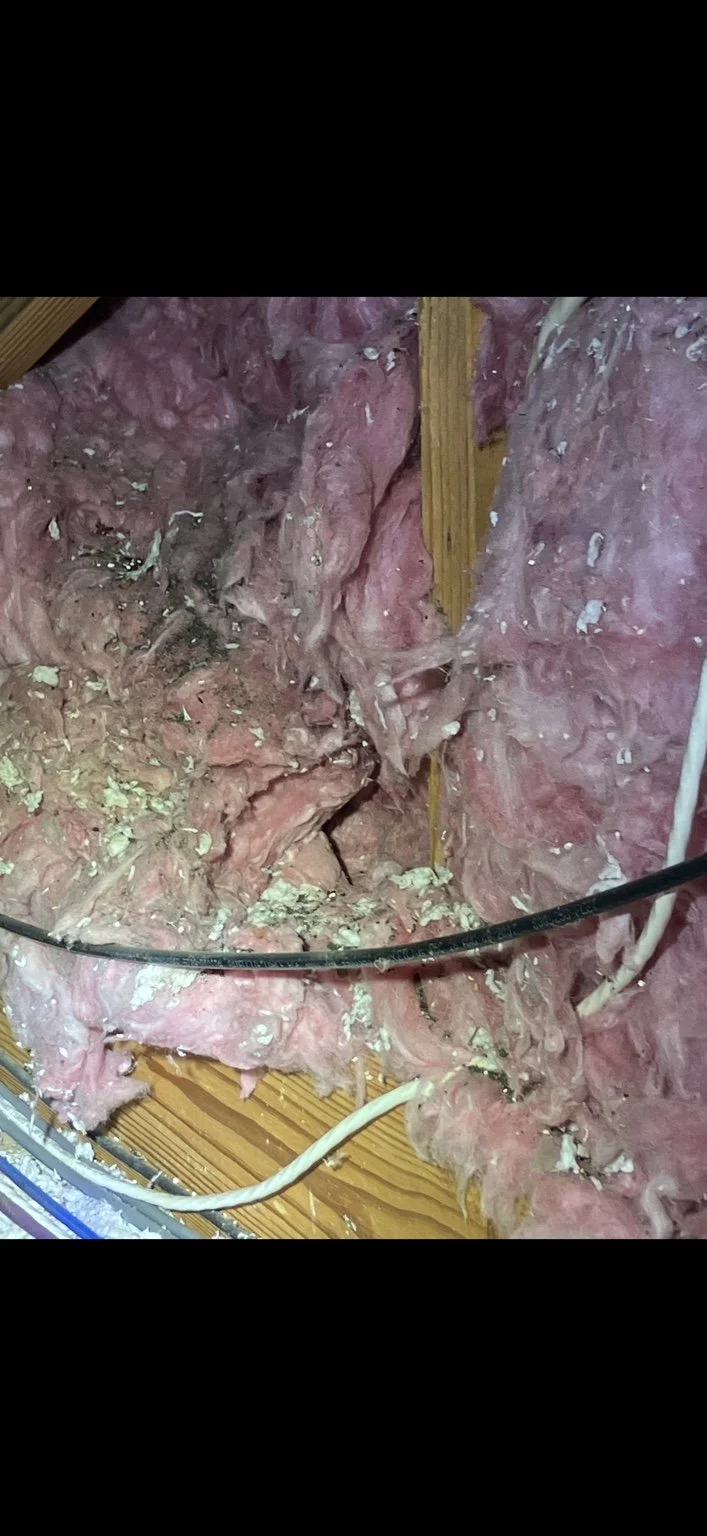 Unfinished attic insulation installation showing pink fiberglass insulation, wooden beams, and electrical wires on a wooden floor.