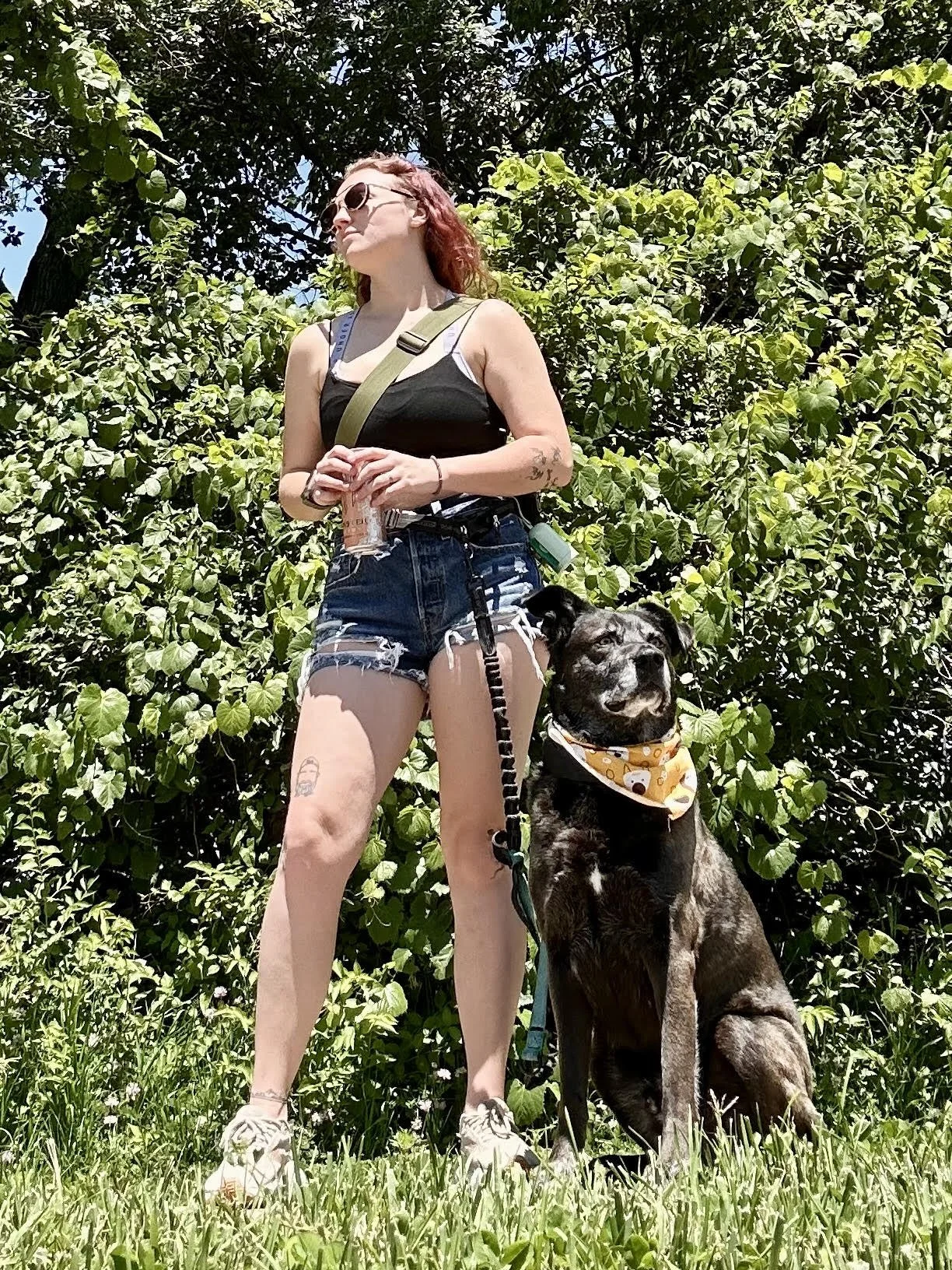 A woman with red hair, sunglasses, tattoos, wearing a black tank top and jean shorts, standing with a large black and gray dog wearing a bandana in green grass with green trees in the background on a sunny day.