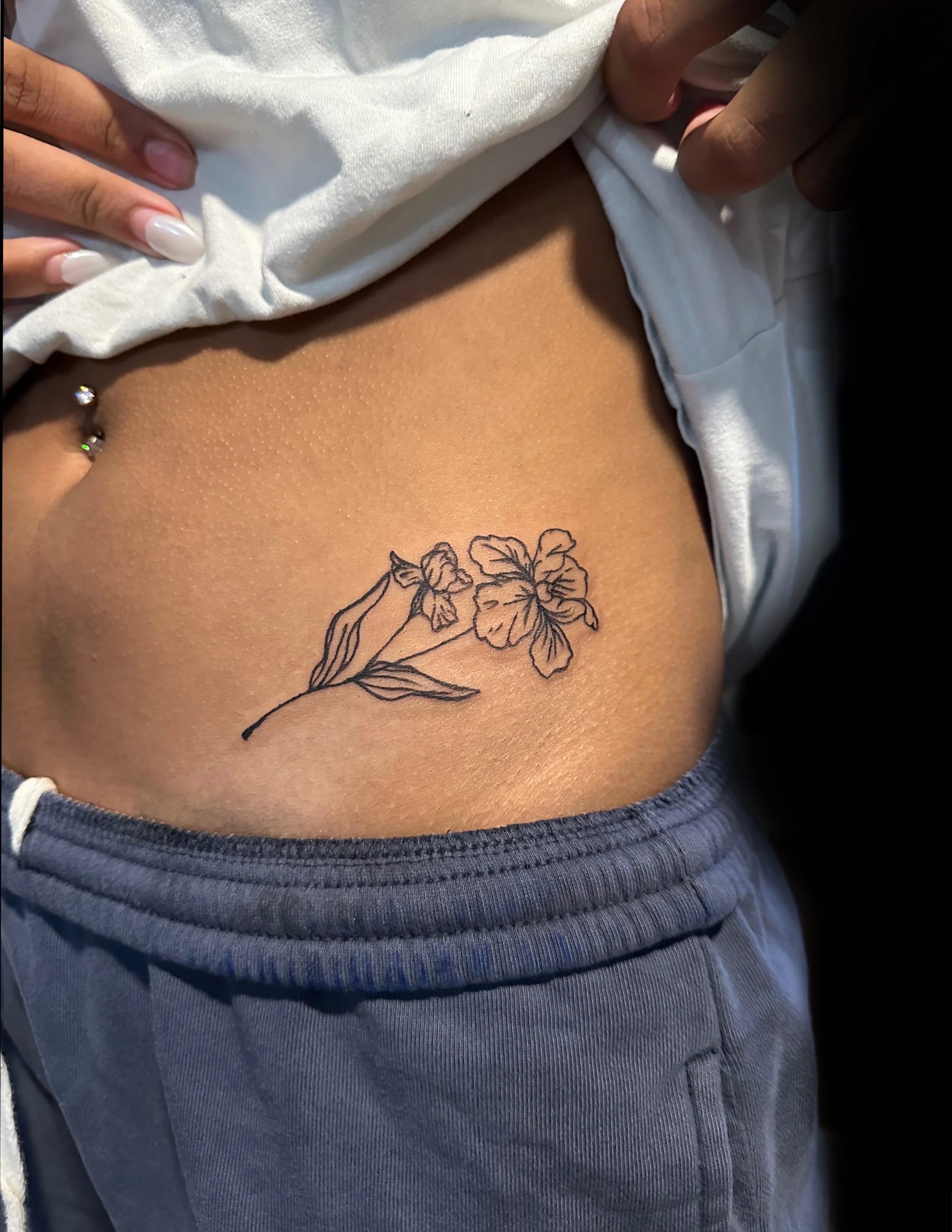 Close-up of a person's hip with a floral tattoo, partially covered by a white shirt and blue jeans.