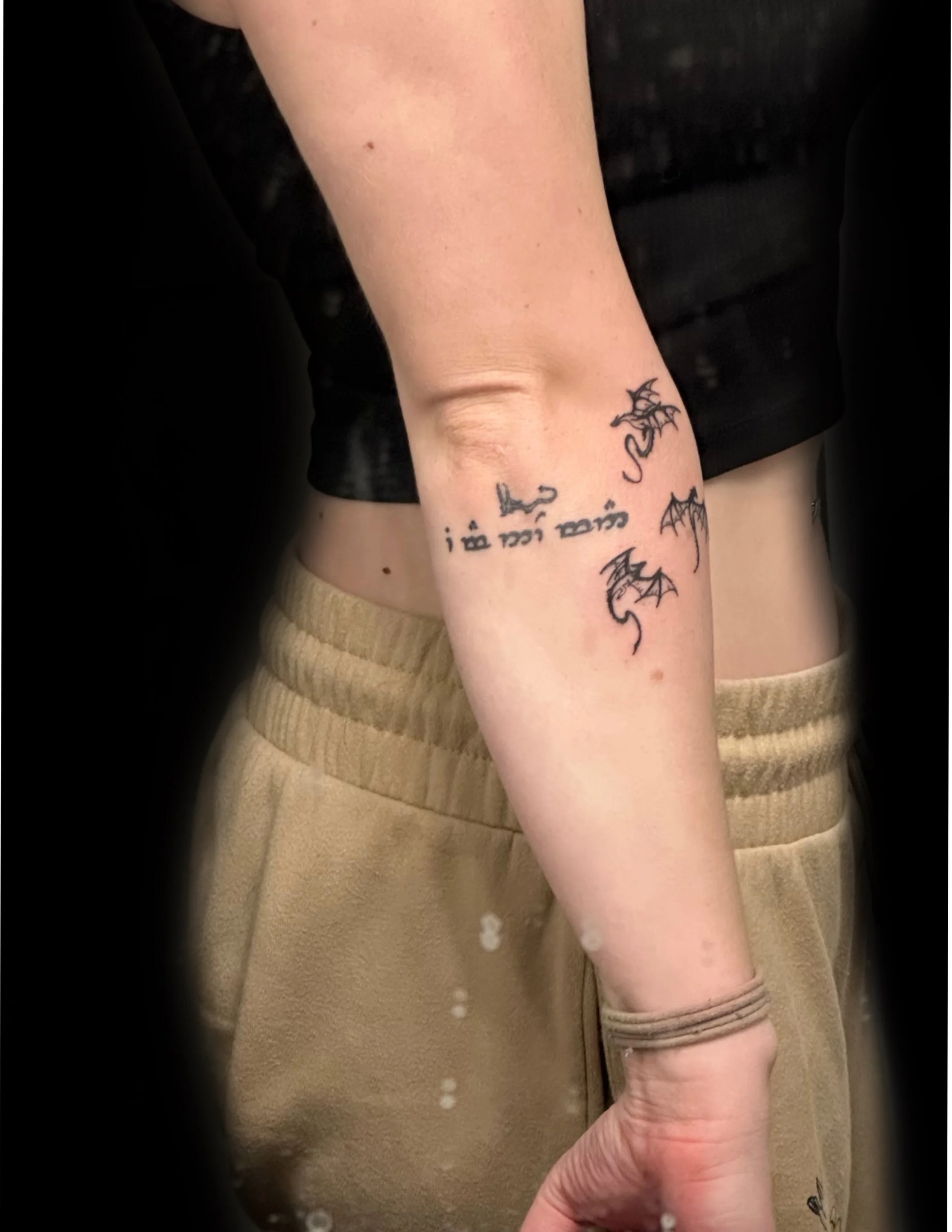 A person's arm with multiple small tattoos, including 3 dragons, the hobbit original drawing of a dragon, and an elfish phrase, dressed in beige pants and a beige shirt, against a dark background.