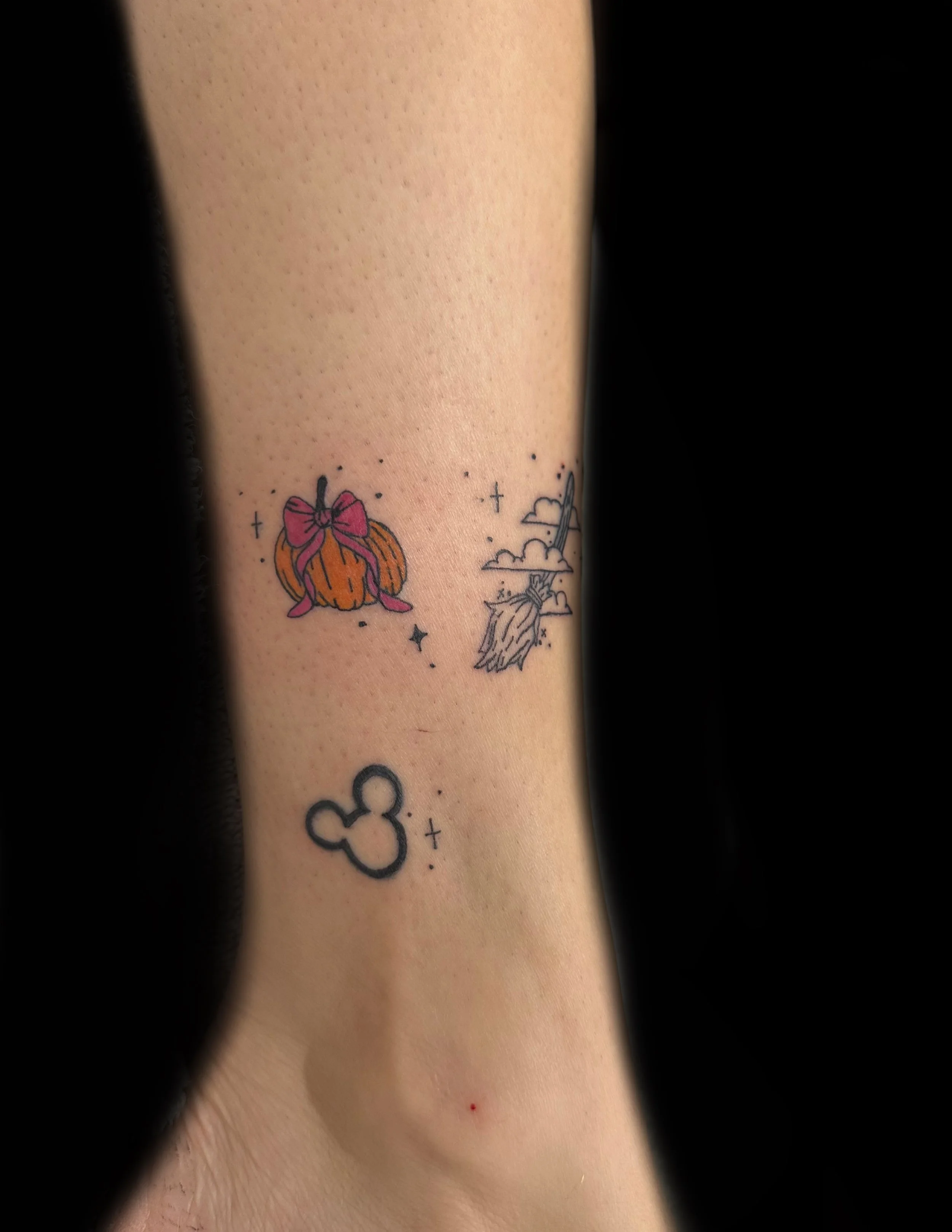 Close-up of a person's forearm with a small tattoo of a pumpkin with a pink bow, a witches broom, stars, and Mickey Mouse head outline.