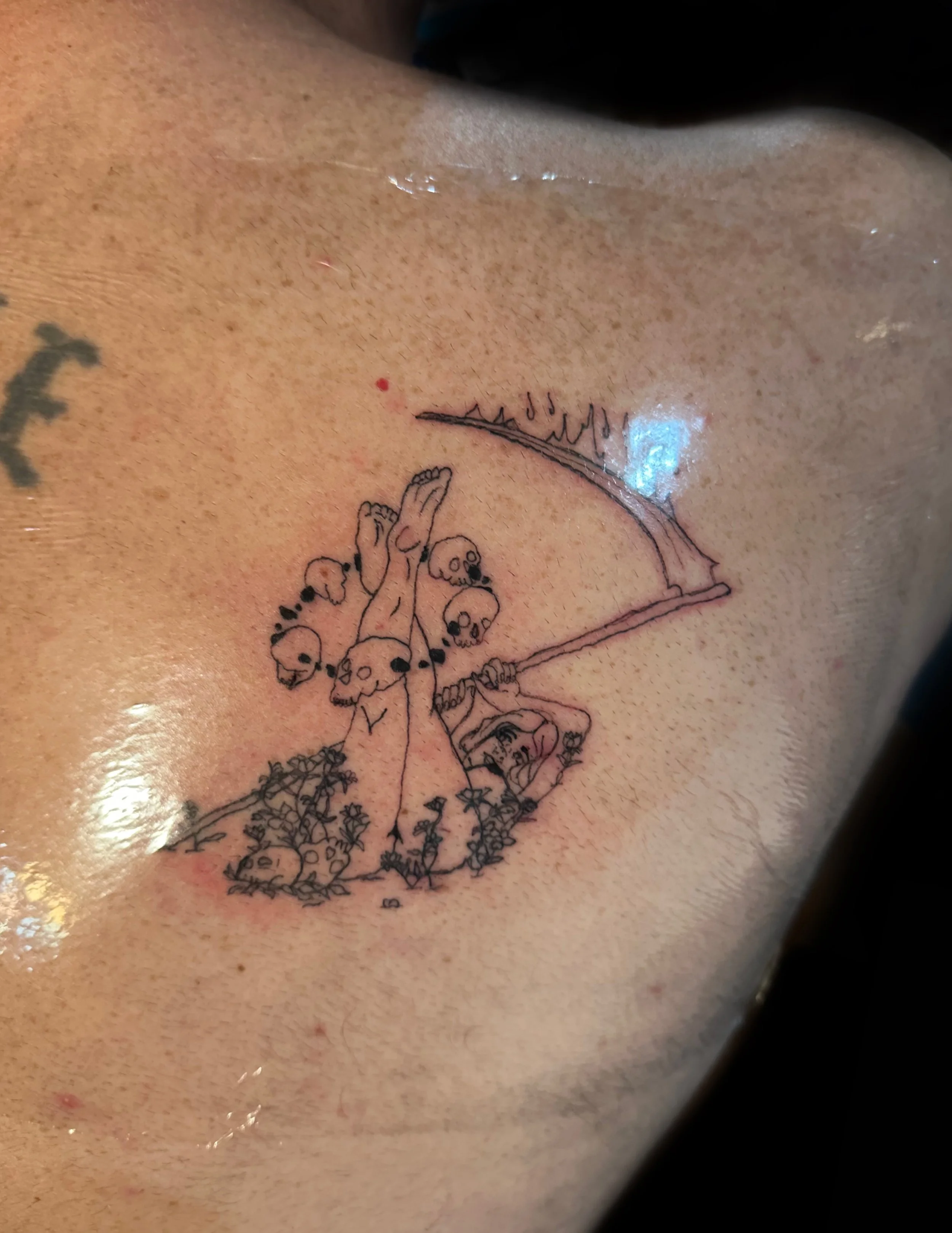 Tattoo of a naked women laying on her back in a field with skulls and flowers holding a scythe with her feet in the air.