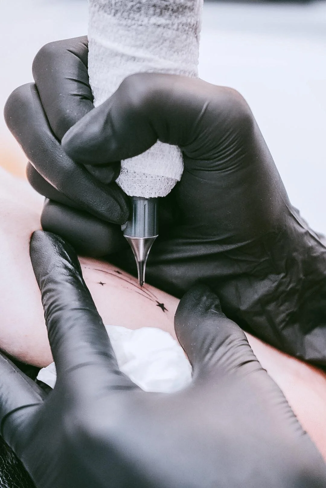 A tattoo artist is creating a small tattoo on a person’s skin using a tattoo gun. The artist is wearing black gloves and the background is blurred.