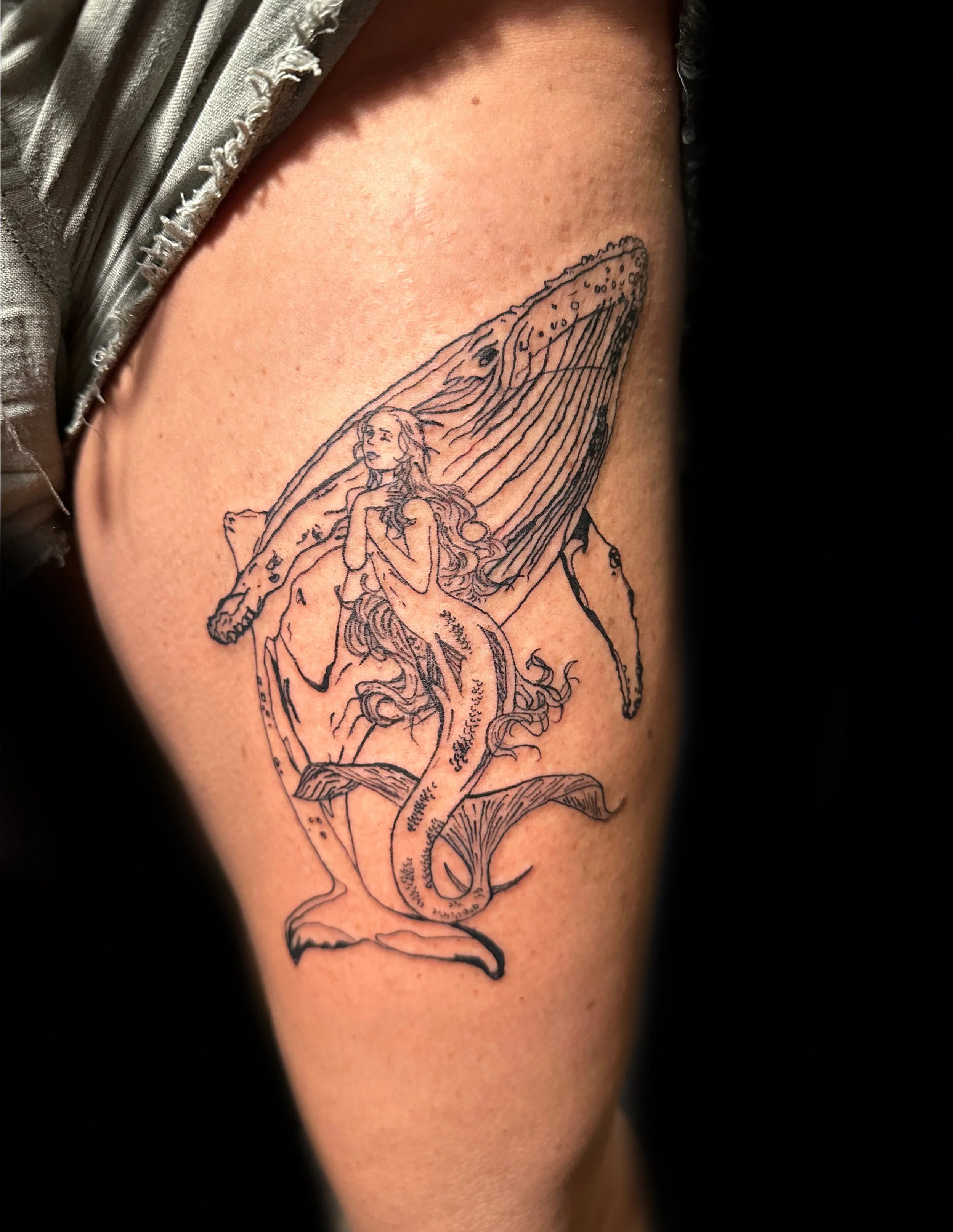 Tattoo of a mermaid in front of a whale on a person's upper arm.
