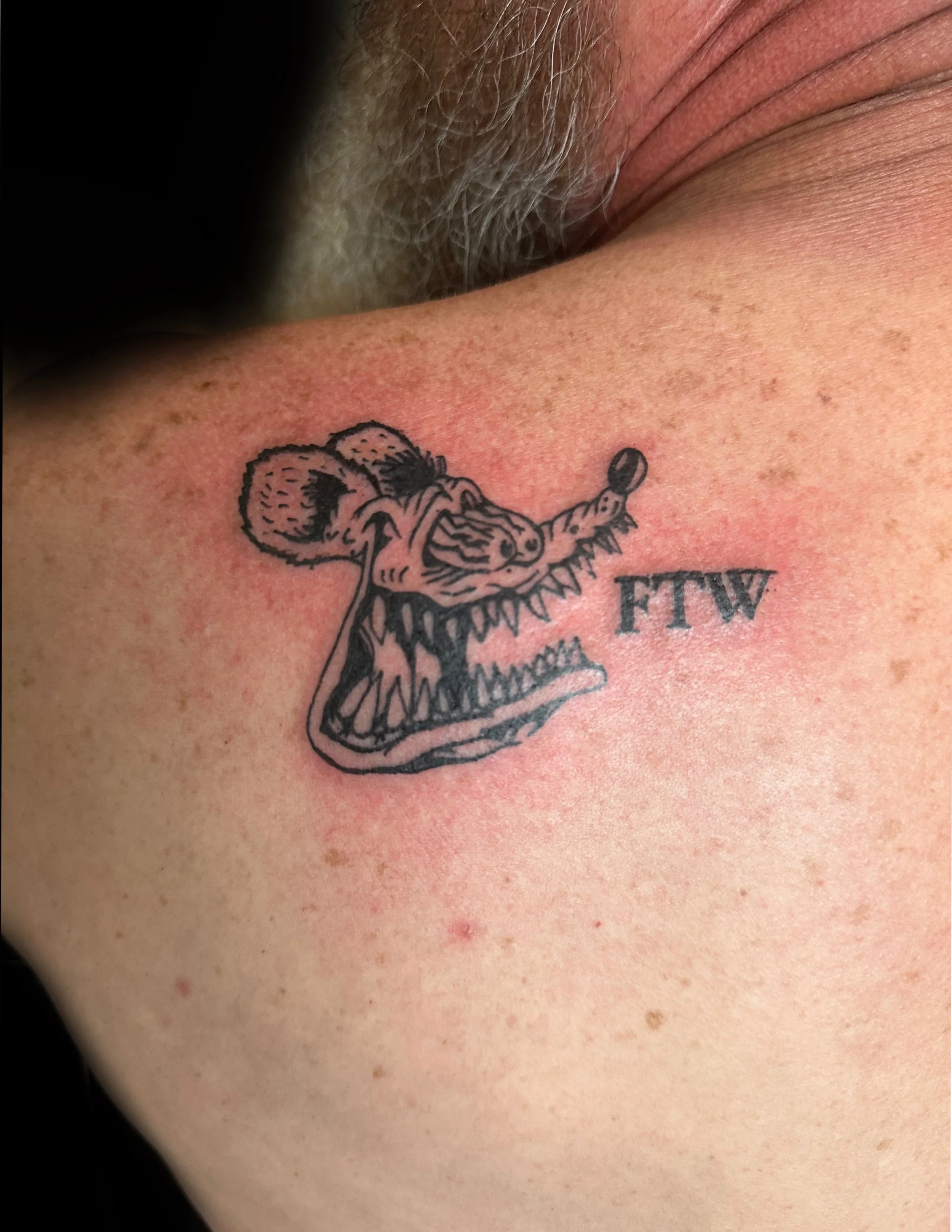 Close-up of a tattoo of a Rat Fink; a cartoonish, open-mouthed rat on human skin, with visible freckles and hair in the background.