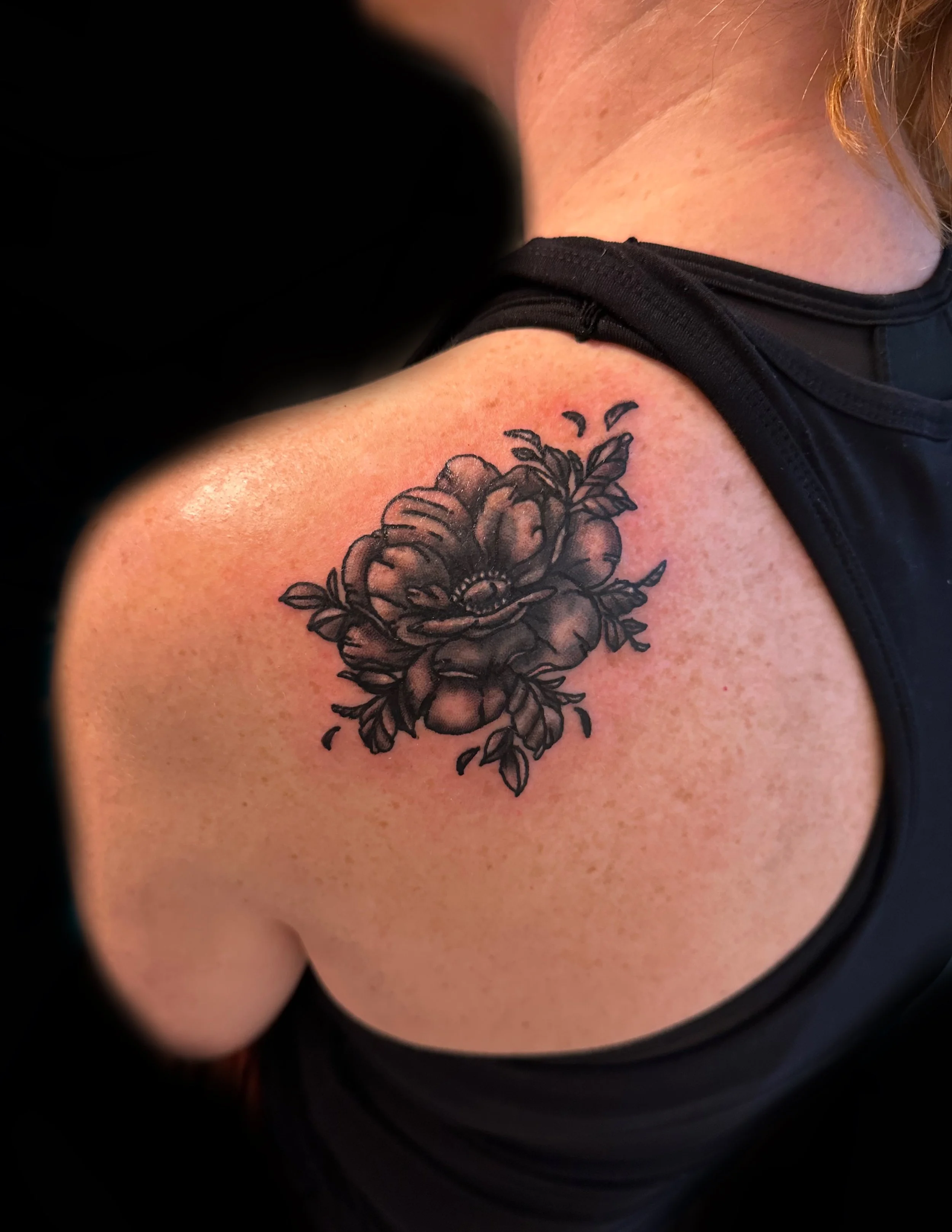 A person's upper arm with a black tattoo of a flower, with the person wearing a black sleeveless top.