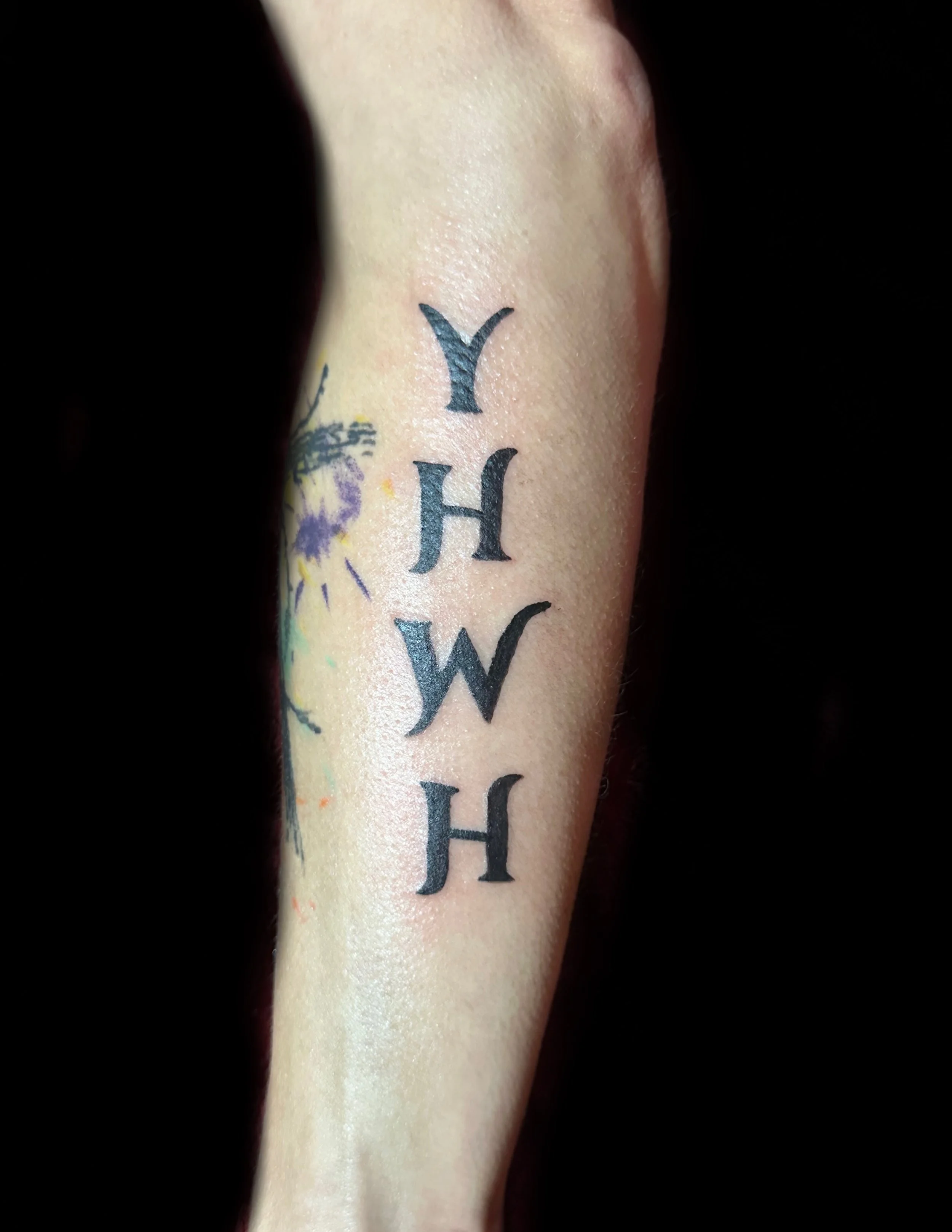 Close-up of a tattoo on skin with black ink lettering spelling 'YHWH', surrounded by black background.
