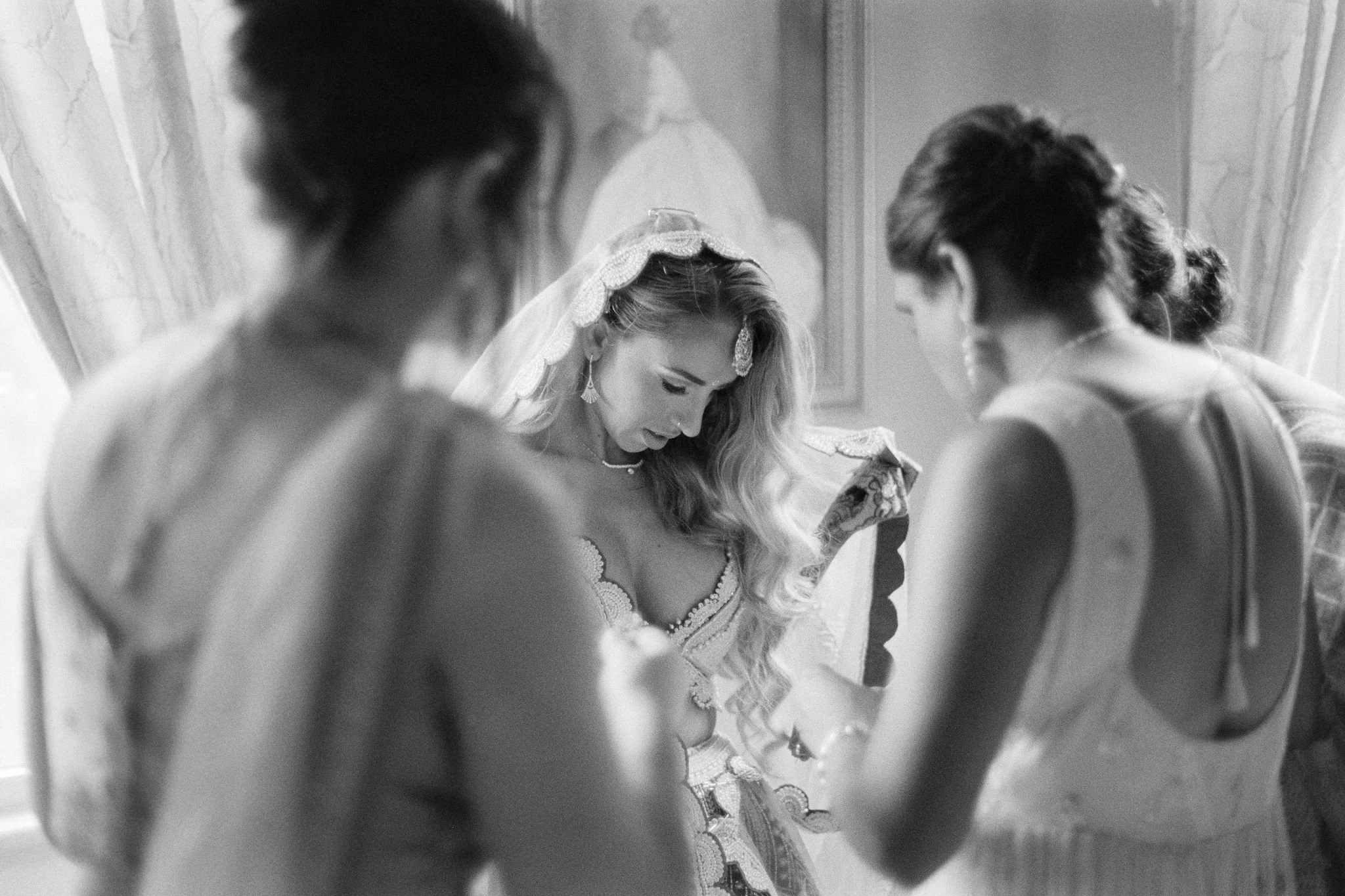A bride with long wavy hair and a veil decorated with lace, surrounded by bridesmaids in dresses, standing in a room with ornate walls, looking down thoughtfully. Shot on black and white film.