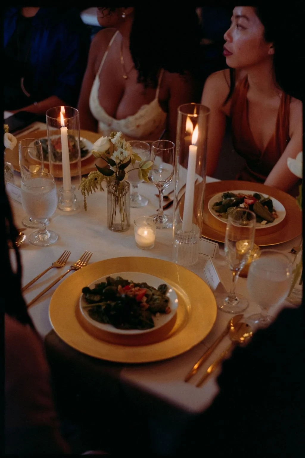 A formal dinner table set with plates of food, wine glasses, lit candles, and floral centerpieces, with women seated around it, dressed elegantly.