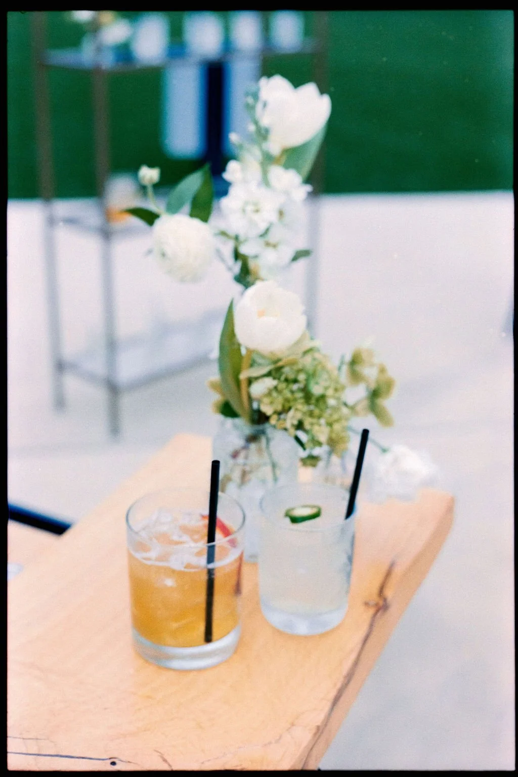 A wooden table with two glasses of cocktails, one orange and one clear with cucumber, in front of a white vase with white flowers and green leaves, set against a blurred background. Shot on Portra 400.