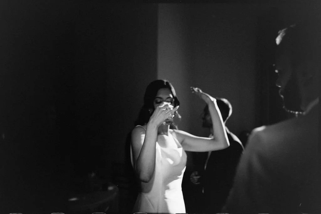 A woman in a white dress drinking from a glass at a social gathering, with two other women in the background, in a dark indoor setting.