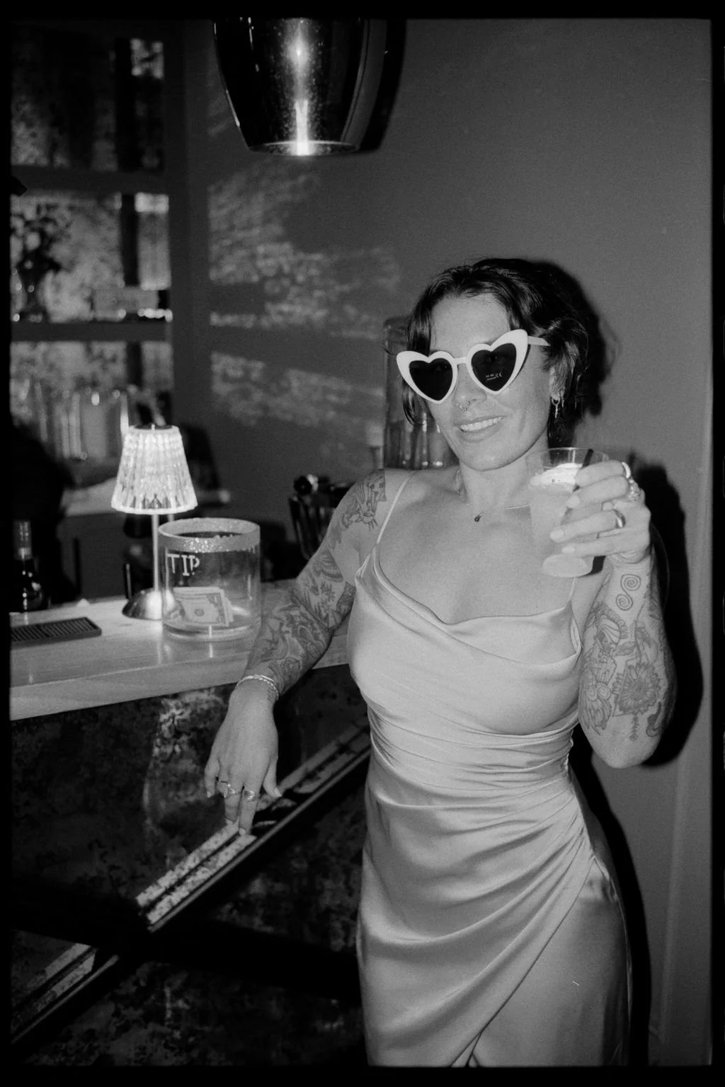 Woman with tattoos wearing heart-shaped sunglasses, holding a cocktail, smiling at a party or celebration.