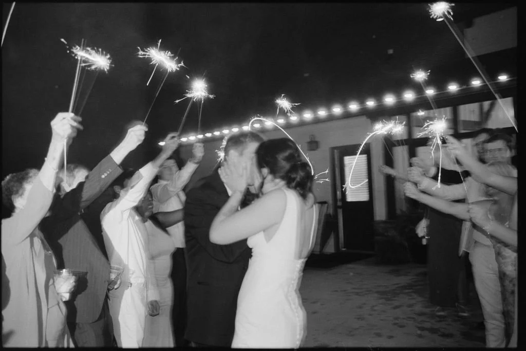 A black and white photo of a wedding celebration with a bride and groom kissing, surrounded by guests holding sparklers in an outdoor setting at night.