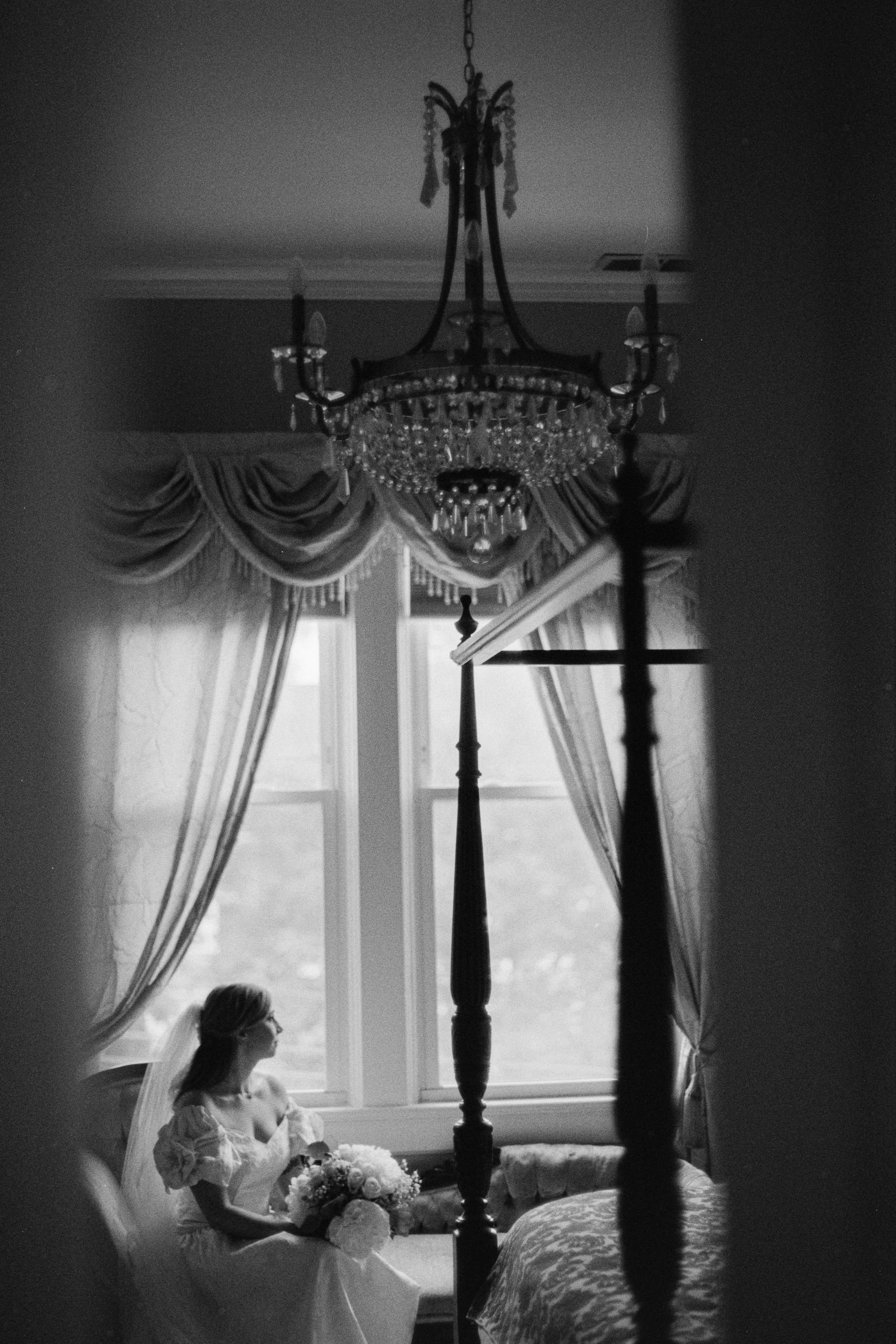 A bride sitting by a window in her wedding dress holding a bouquet, with a chandelier hanging above and draped curtains in the background.