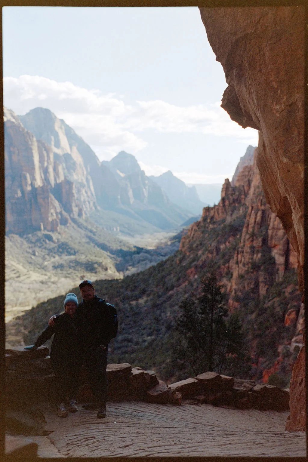 Two people, a man and a woman, standing together on a rocky ledge inside a canyon with towering cliffs and mountains in the background.
