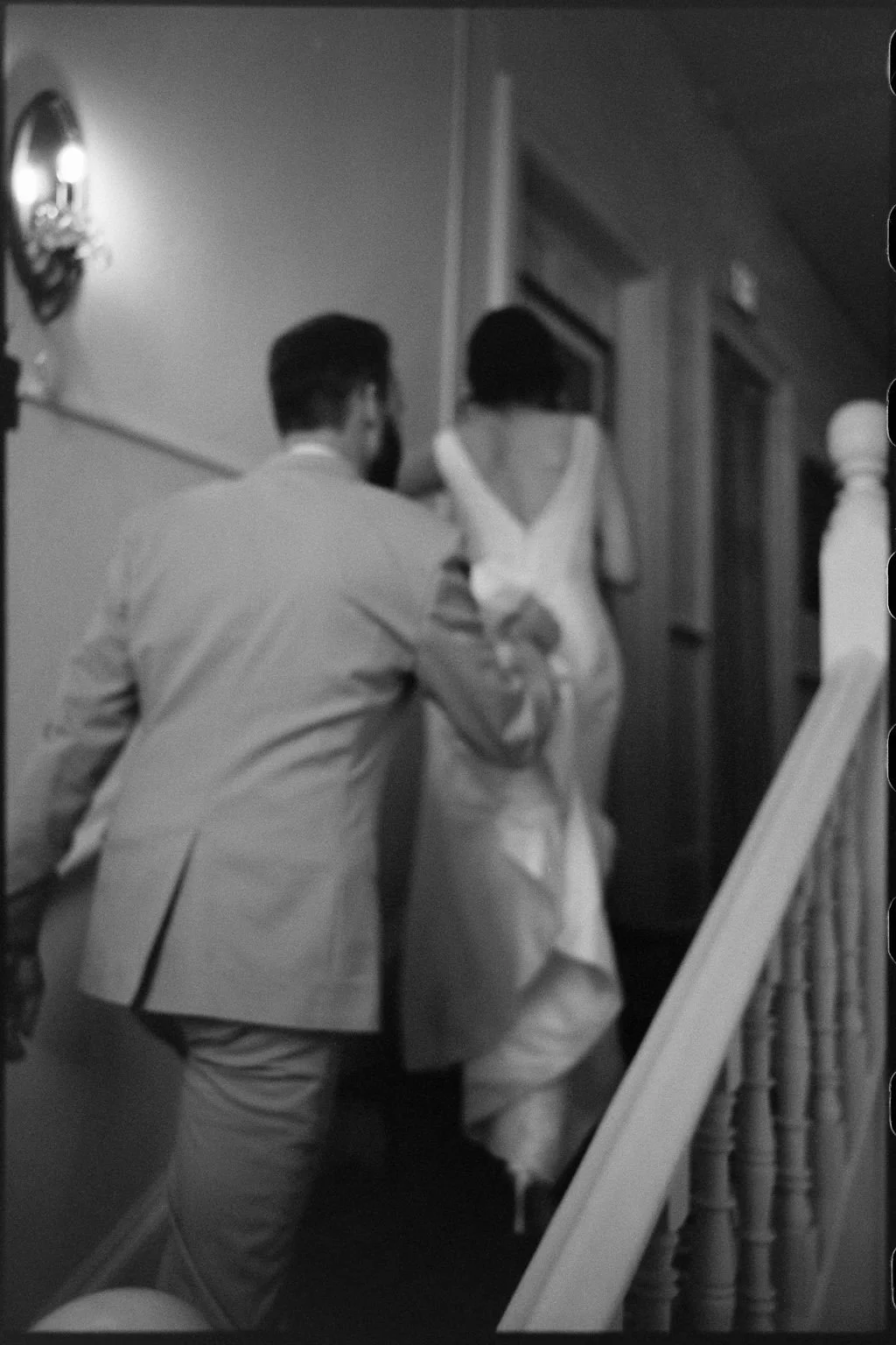 A black-and-white photo depicts a man assisting a woman in a dress as she ascends a staircase.