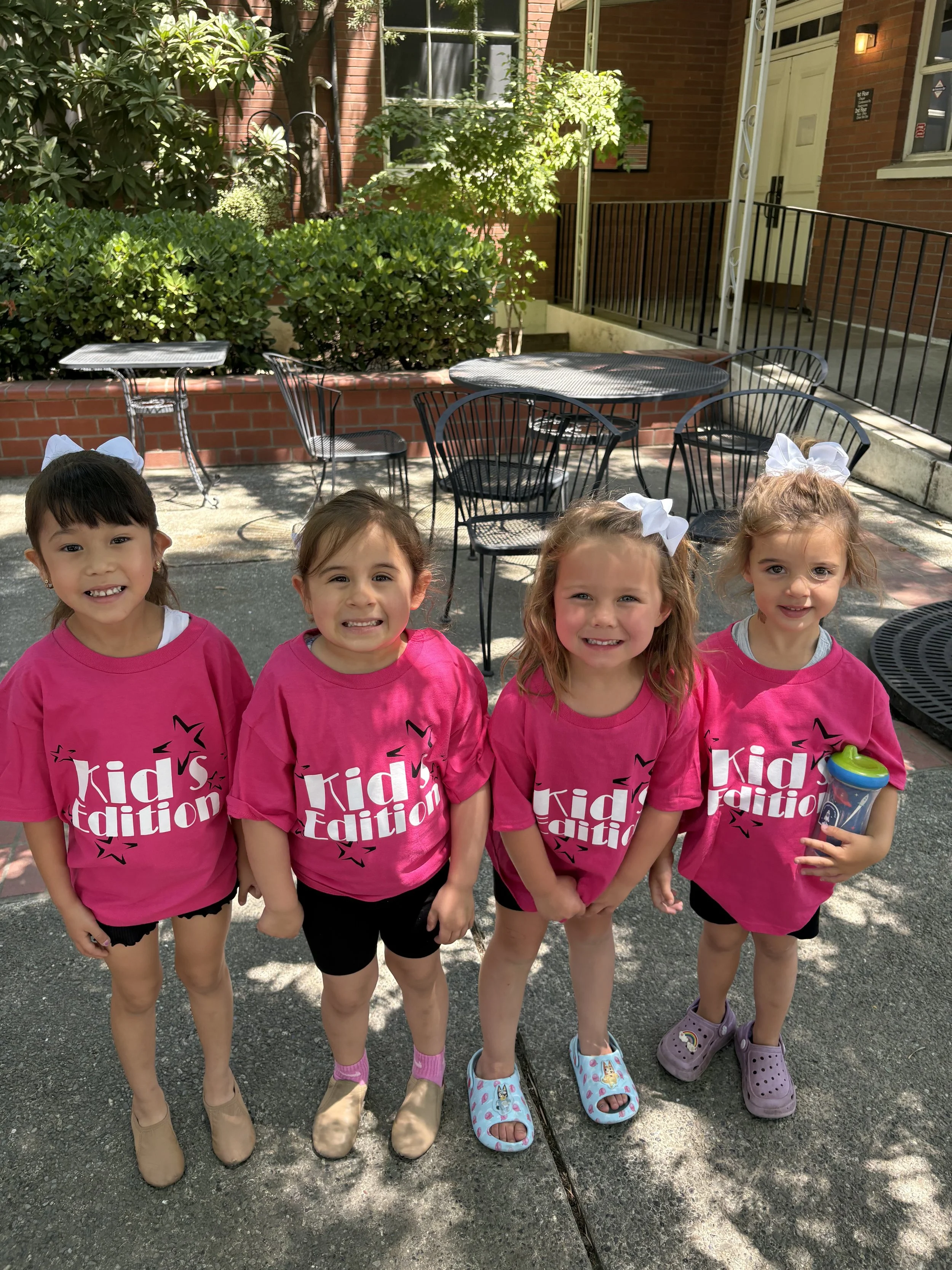 Four young girls standing outdoors in front of a low brick wall with green bushes and a building with red brick walls and black metal railings behind them. They are smiling and wearing bright pink t-shirts with the text 'Kids Edition' printed on them, and wearing different types of shoes, including Crocs and sandals. Two girls have large white bows in their hair.
