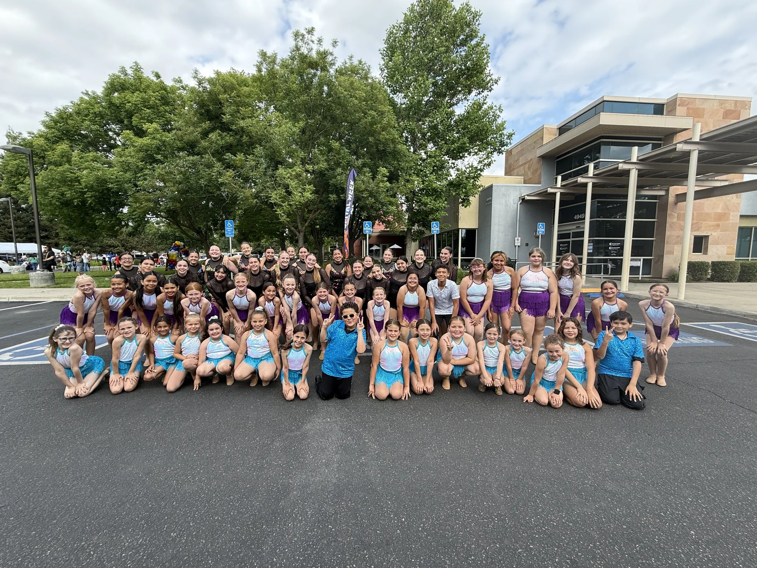 Group of young dancers in colorful costumes, posing outdoors in a parking lot in front of trees and a modern building.