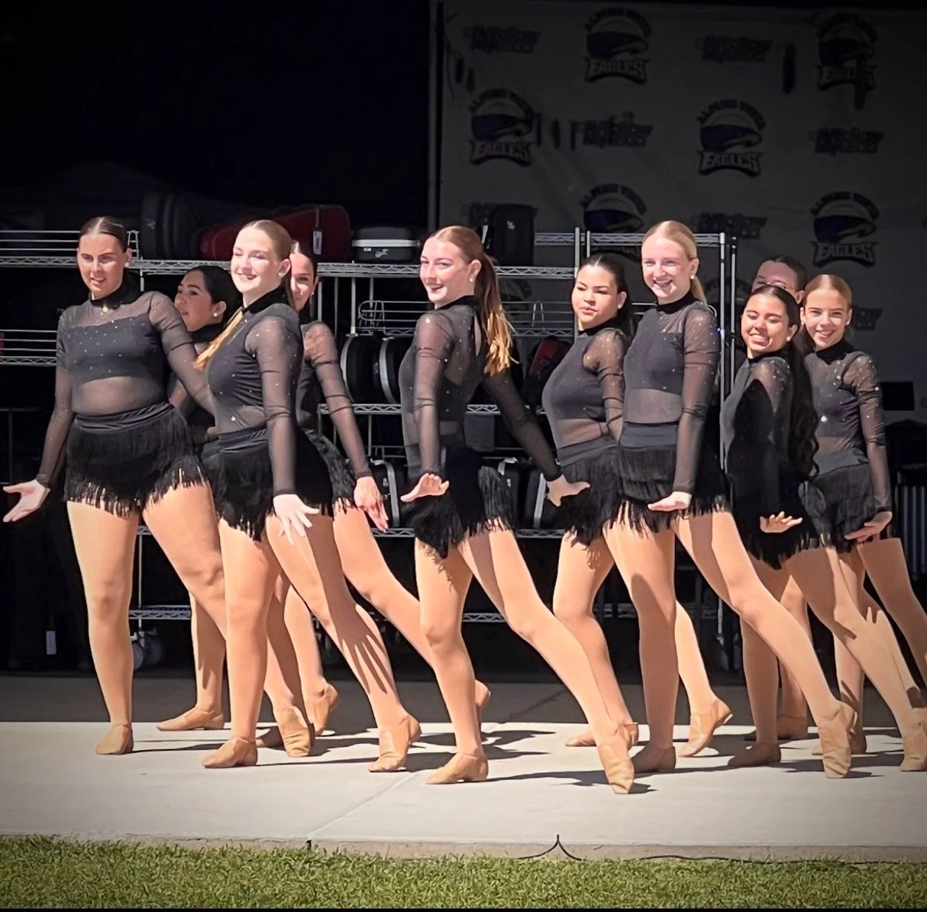 Dance team practicing on stage in black costumes with sheer long sleeves and fringe skirts, performing synchronized dance moves.