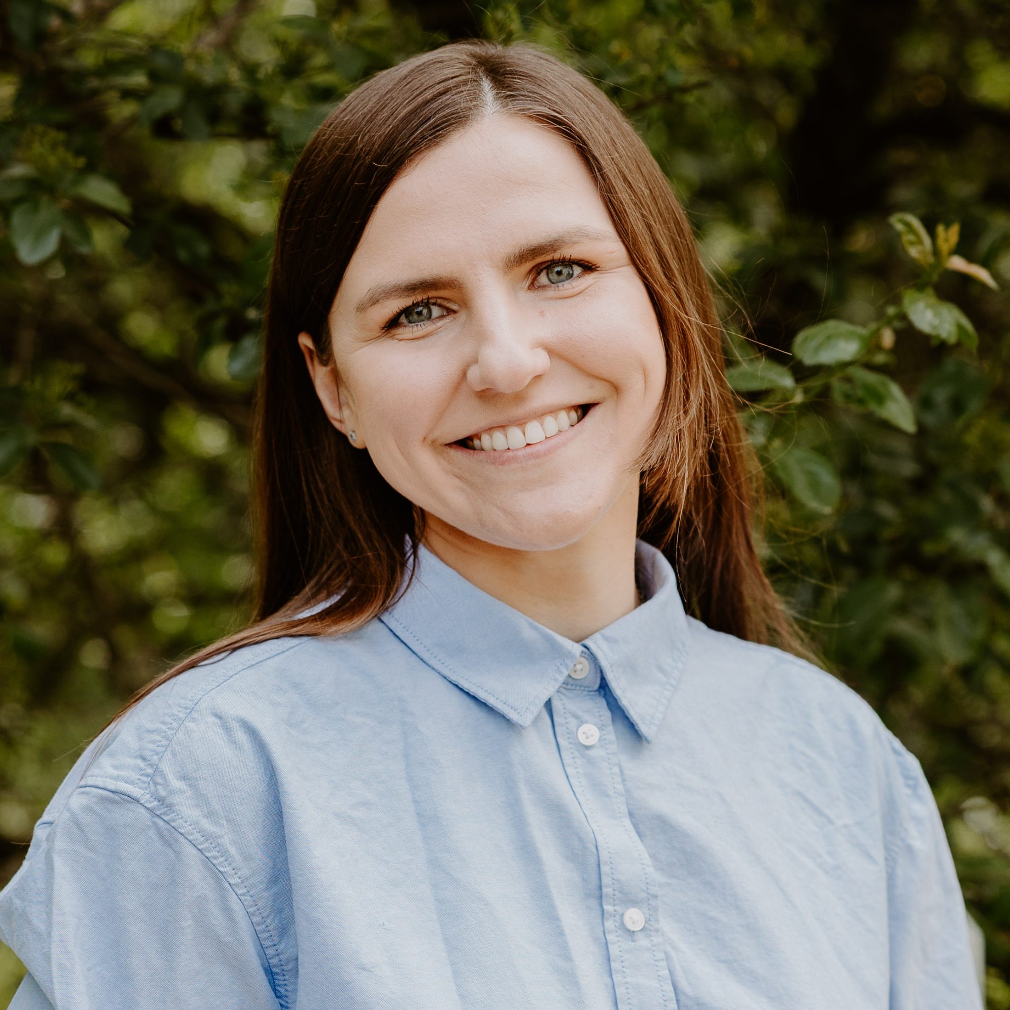 A smiling woman with long brown hair, wearing a black denim jacket and a blue top, outdoors with green foliage in the background.
