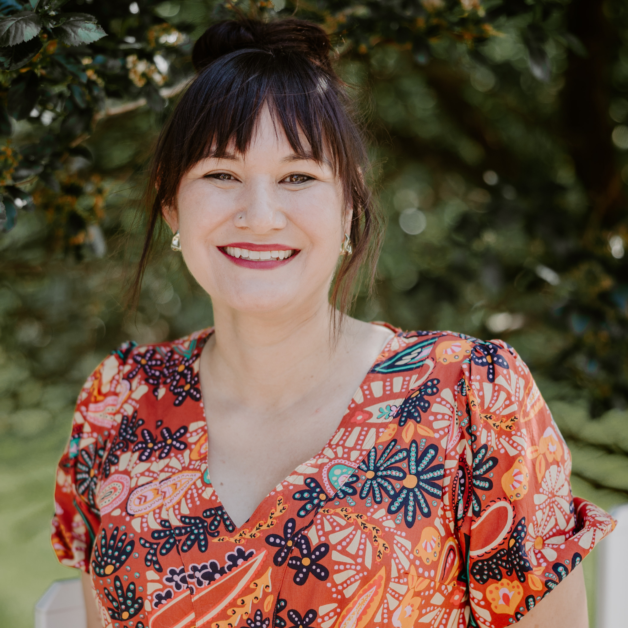 A woman with shoulder-length dark brown hair, wearing a floral blouse, smiling outdoors with green trees in the background.