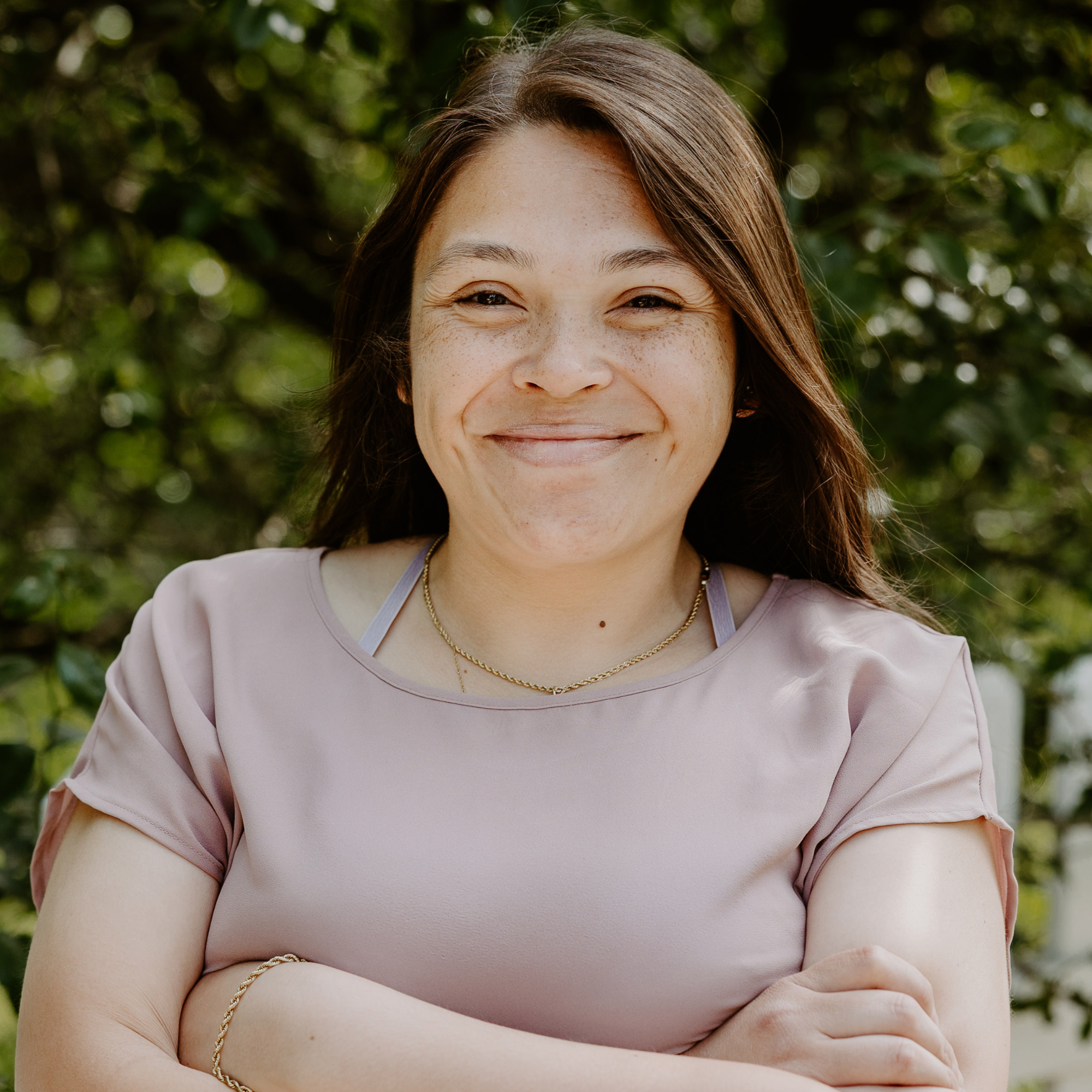 A woman with long dark hair smiling outdoors with green leaves and trees in the background.