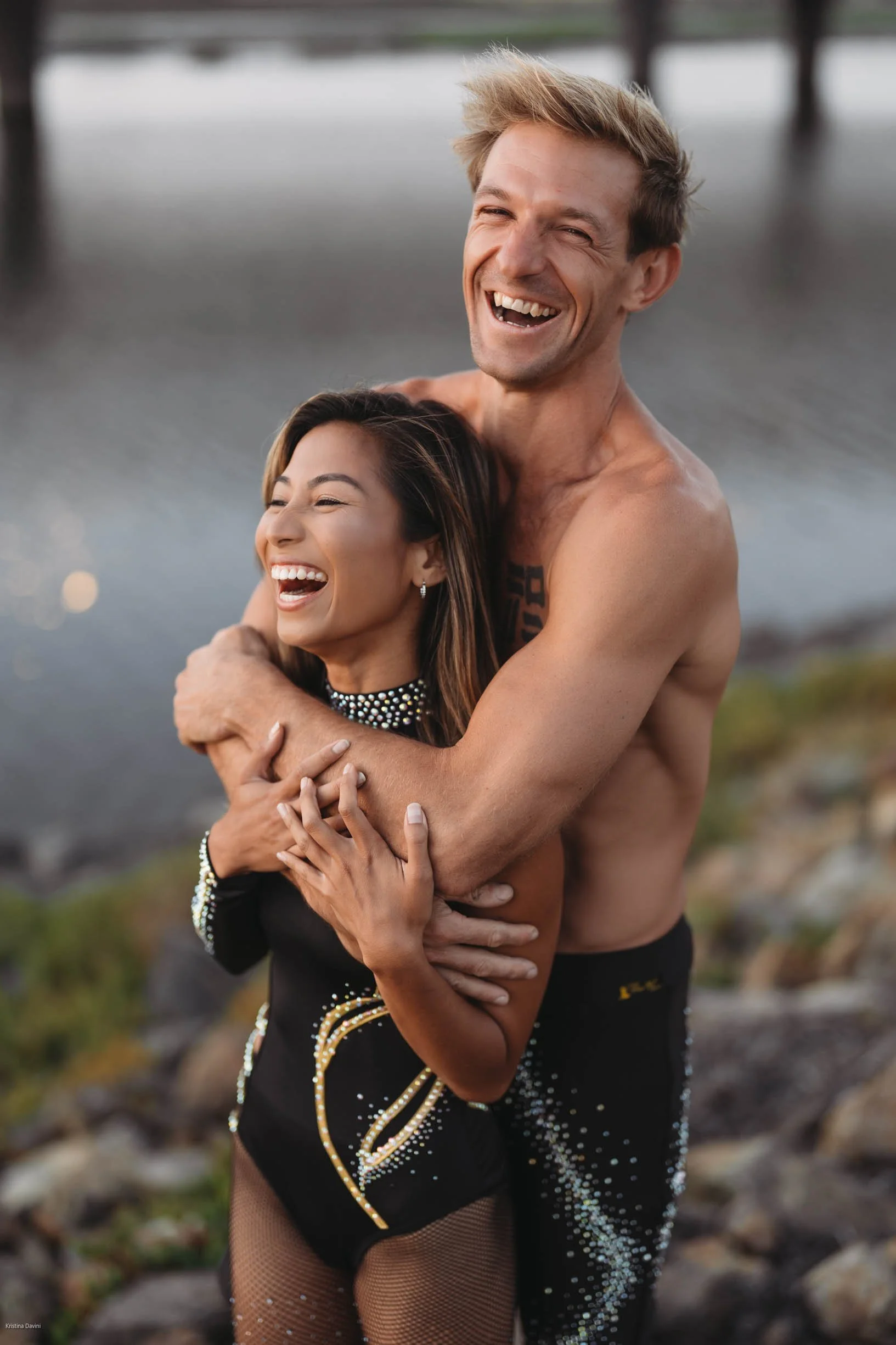 A smiling man with short, styled blond hair hugging a smiling woman with long, dark hair. They are shirtless and outdoors near water, with a pier or dock in the background.
