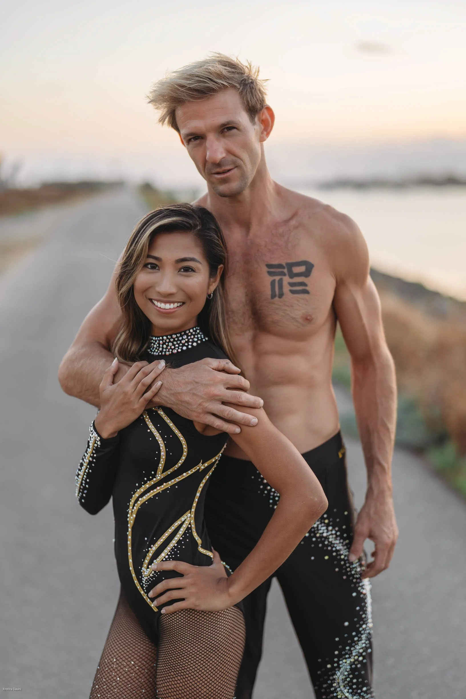 A man and woman standing outdoors near a body of water during sunset. The man is shirtless with a tattoo on his chest, and the woman is smiling, wearing a black dress with gold and silver embellishments and fishnet stockings.