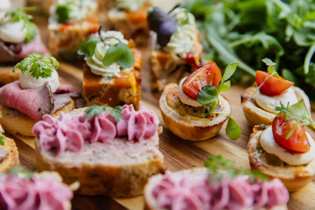 Assorted small bites and canapés topped with vegetables, meats, and herbs on a wooden serving board.
