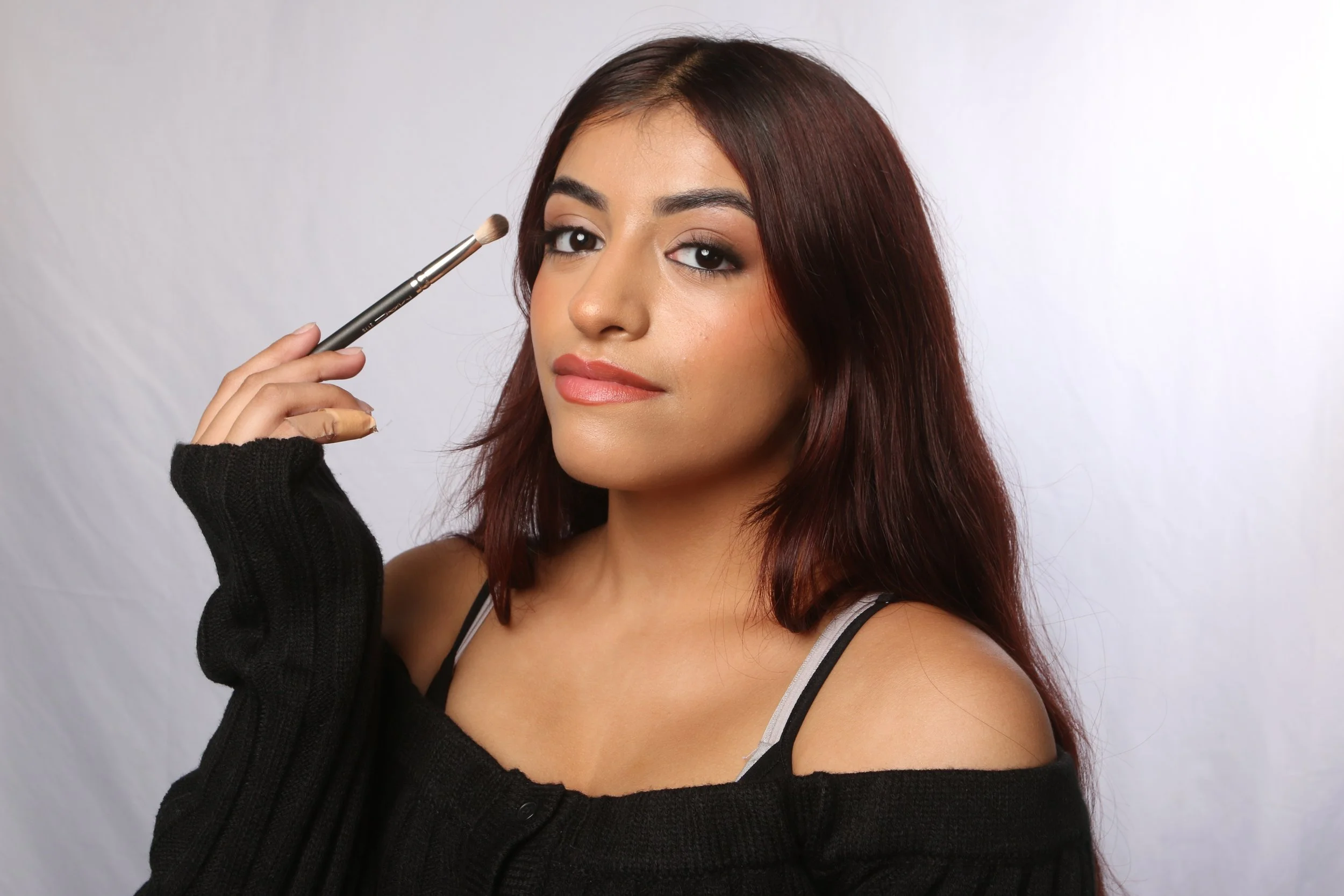 A woman with long reddish-brown hair, makeup, and wearing a black sweater, is holding a makeup brush near her face, against a plain light-colored background.