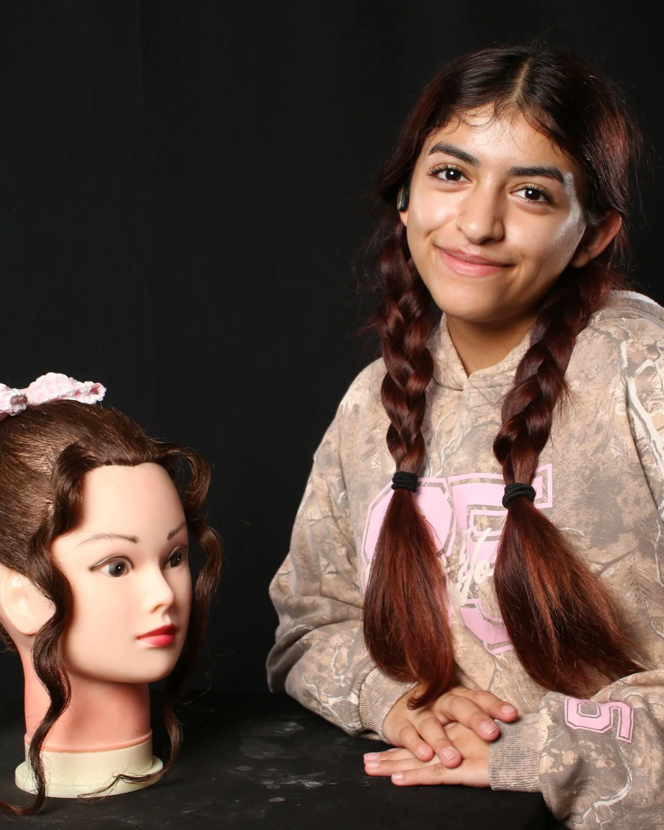 A girl with long, brown braided hair, wearing a beige hoodie with a pink logo, smiles at the camera, sitting next to a mannequin head with long, curly, brown hair and a pink bow, against a black background.