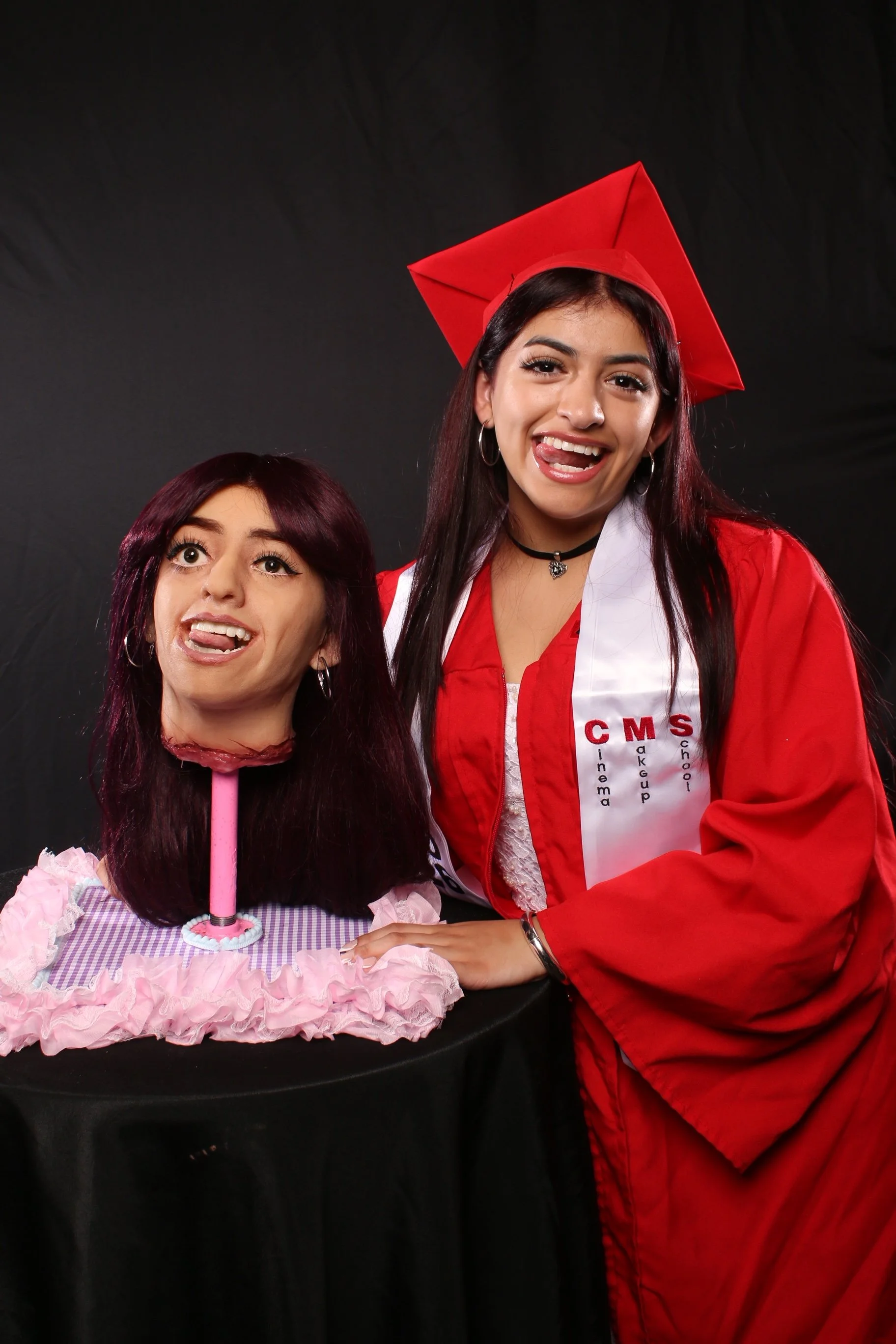 Two young women celebrating graduation, one wearing a red cap and gown, the other with a prop doll. They are smiling and posing against a black background.