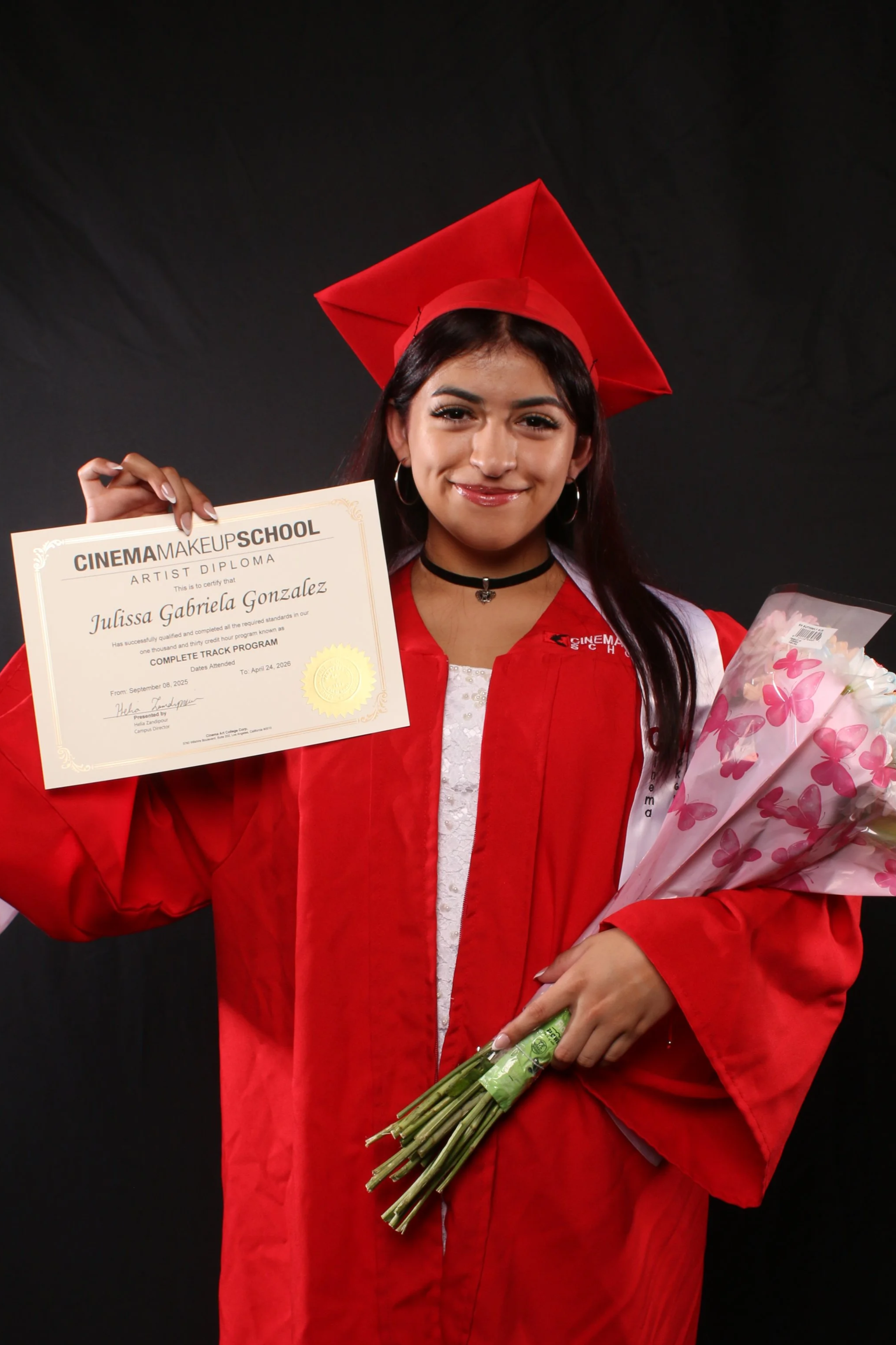 A young woman with long dark hair, wearing a red graduation cap and gown, is smiling and holding a diploma in one hand and a bouquet of pink flowers in the other. The background is black.