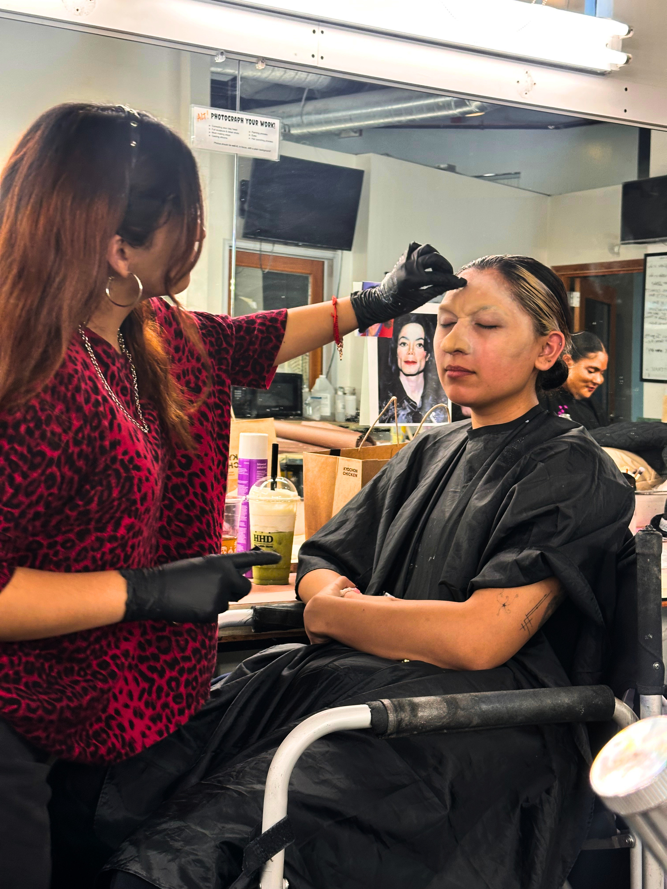 A woman with closed eyes getting her makeup done by a makeup artist at a salon or studio. The woman is seated in chair, wearing a black cape, with blonde hair slicked back. The makeup artist is wearing a red leopard print top and black gloves, applying makeup on the woman's face. In the background, there are posters, a television, and various salon products.