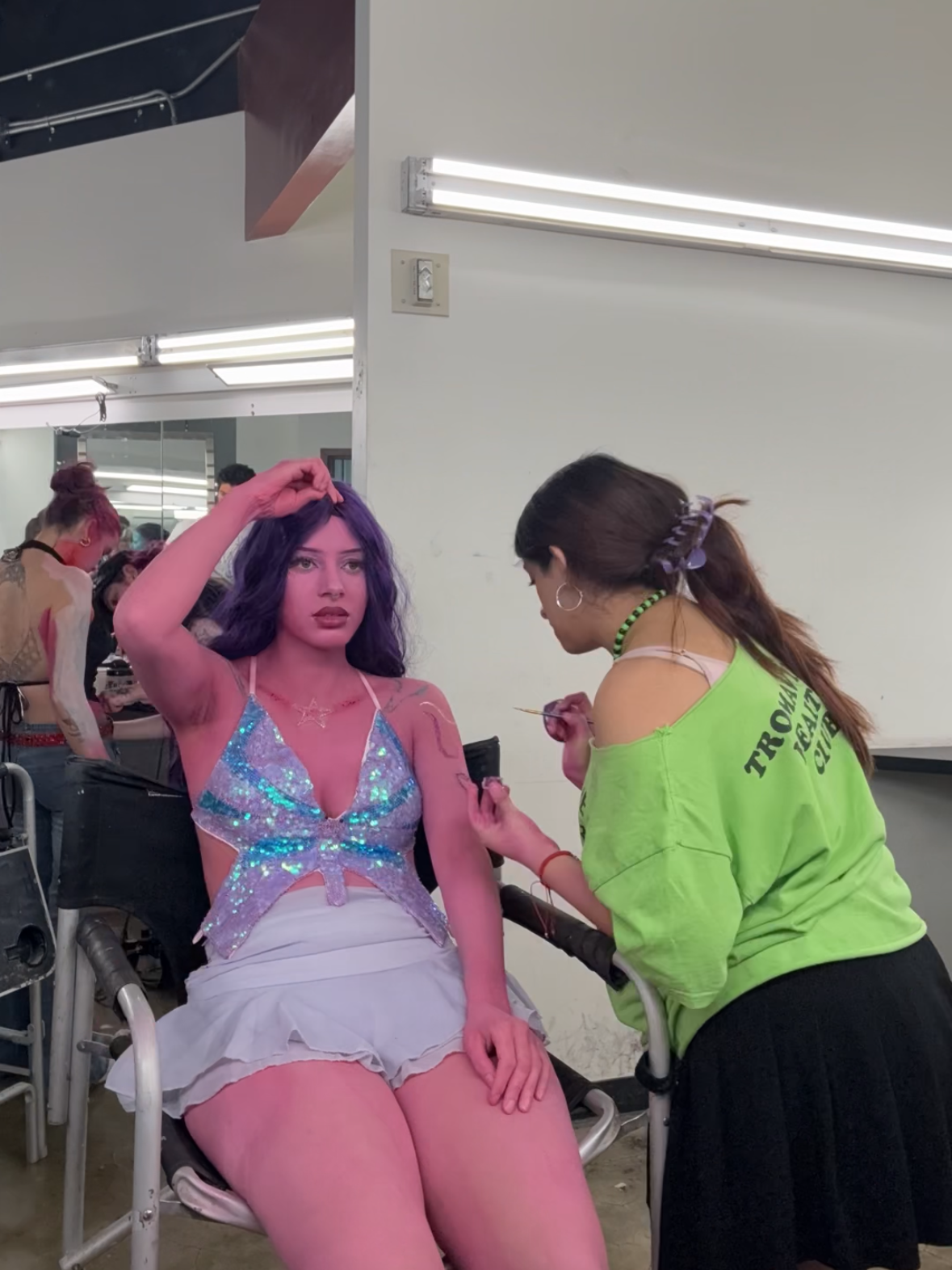 A woman with purple hair sitting on a chair getting her makeup done by a makeup artist in a backstage area.