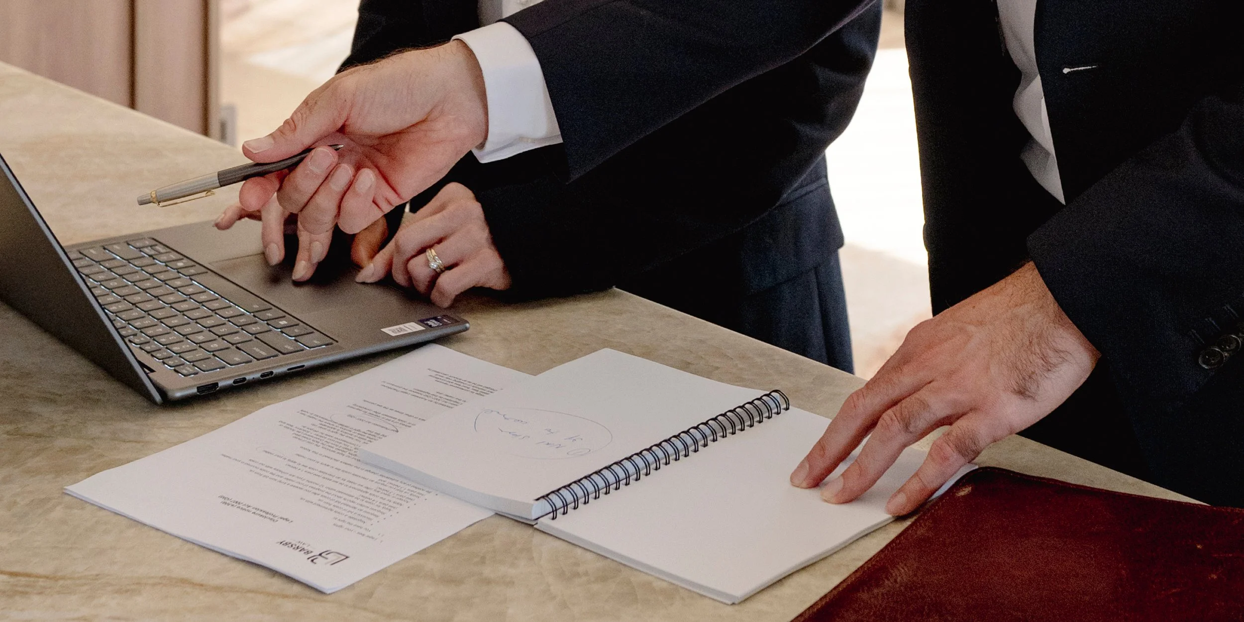 Two individuals in business attire are reviewing documents and working on a laptop at a conference table.