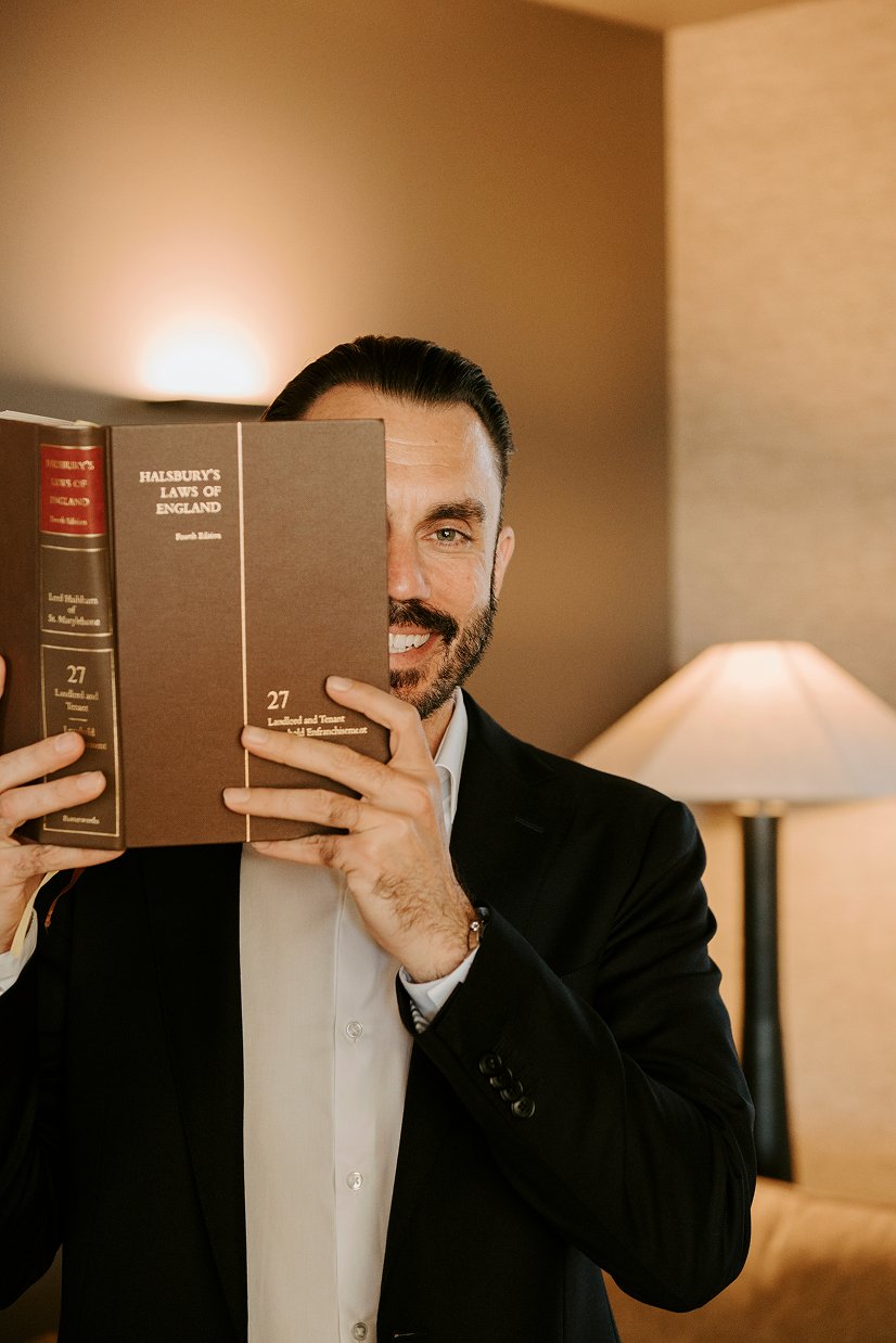 A man in a tuxedo holding a large book titled "Halsbury's Laws of England" partially covering his face, with only one eye and part of his face visible.